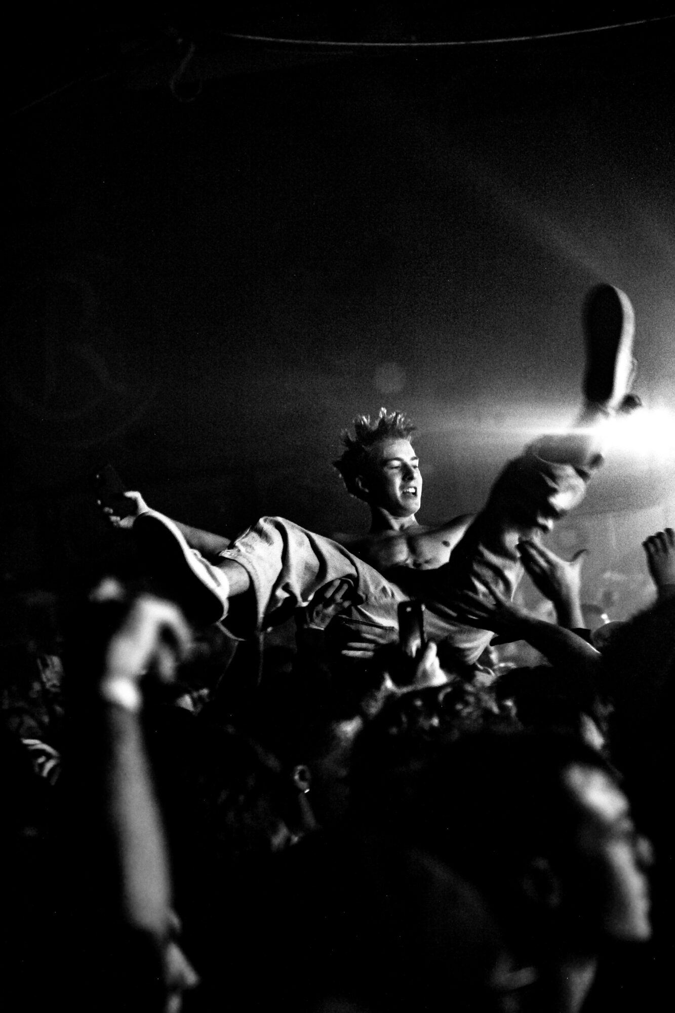 Person crowd surfing at a concert with raised hands and a bright light in the background.
