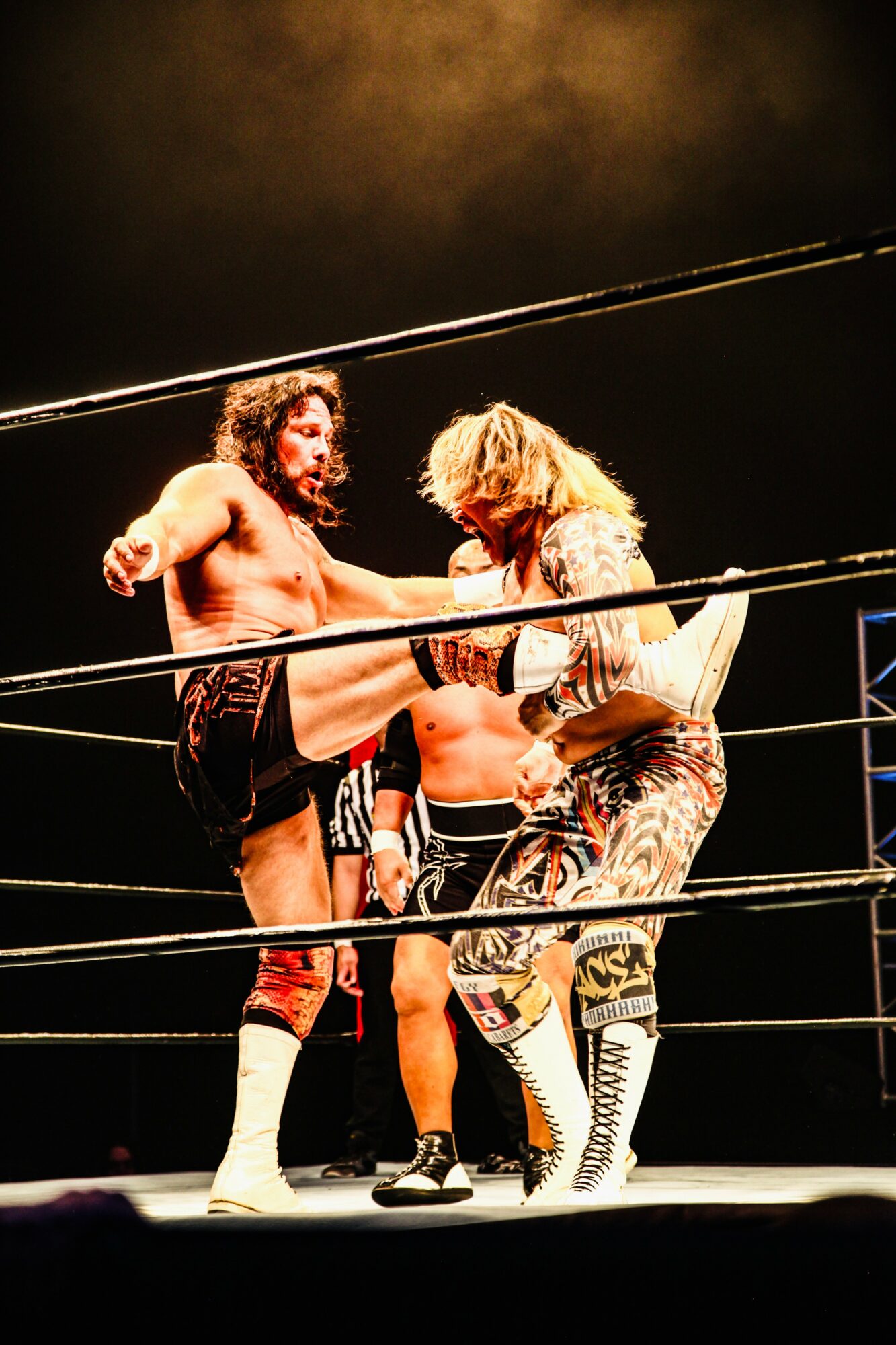 Two wrestlers face off in a ring, one with long dark hair and the other with blonde hair, both in wrestling attire.