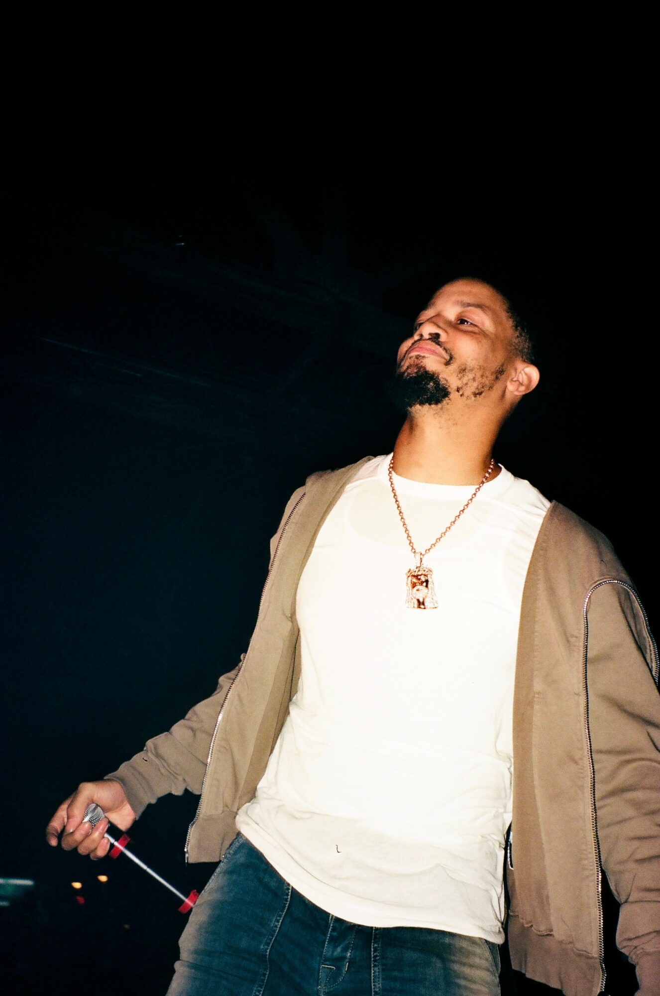 Man with beard and glasses smiling, holding a stick, wearing a white shirt and beige jacket, against a dark background.