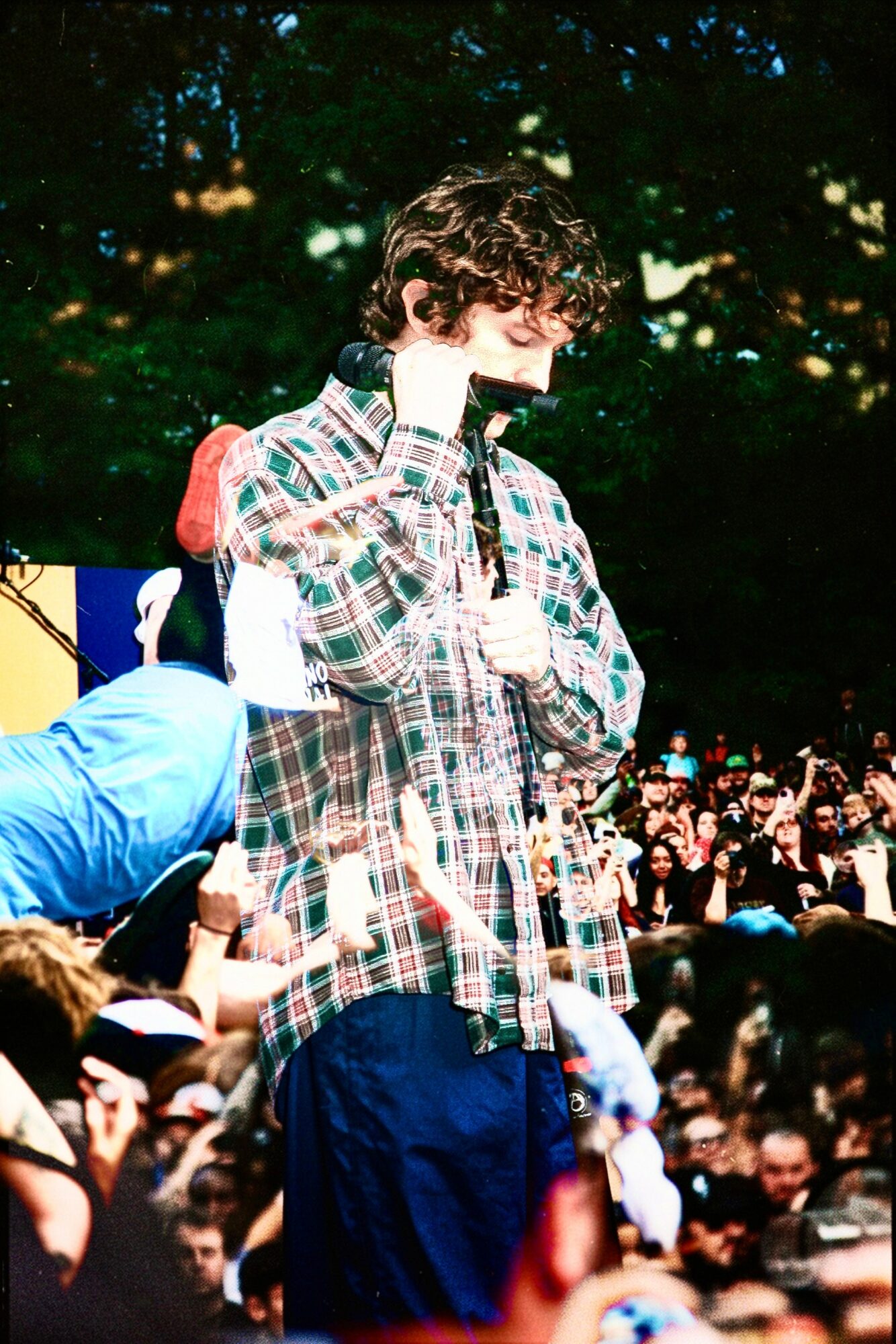 Young person with curly hair wearing a plaid shirt and backpack, surrounded by a crowd outdoors.