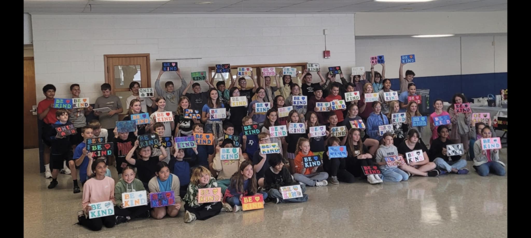 Large group of children and adults in a school hallway holding signs, some sitting and some standing, with lockers in the background.