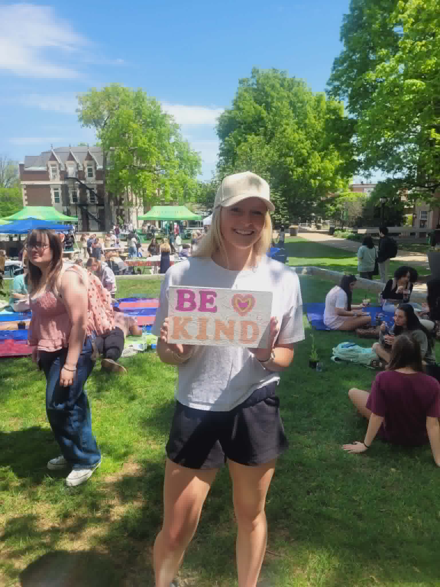 Woman holding a sign that says 'BE KIND' in a park with people, trees, and tents in the background.