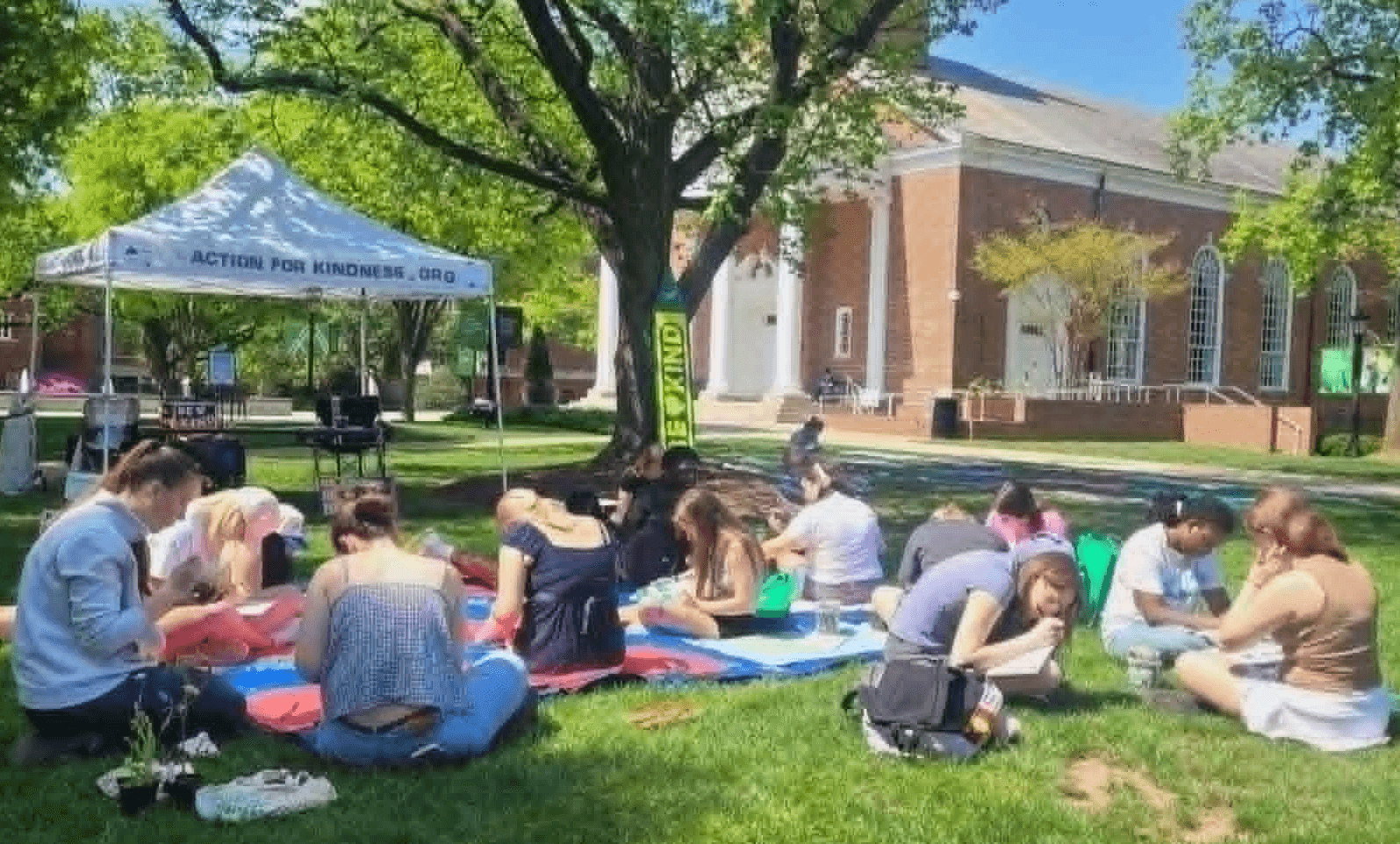 Group of people sitting on blankets and chairs outdoors under a tree, participating in an activity near a building and tent.