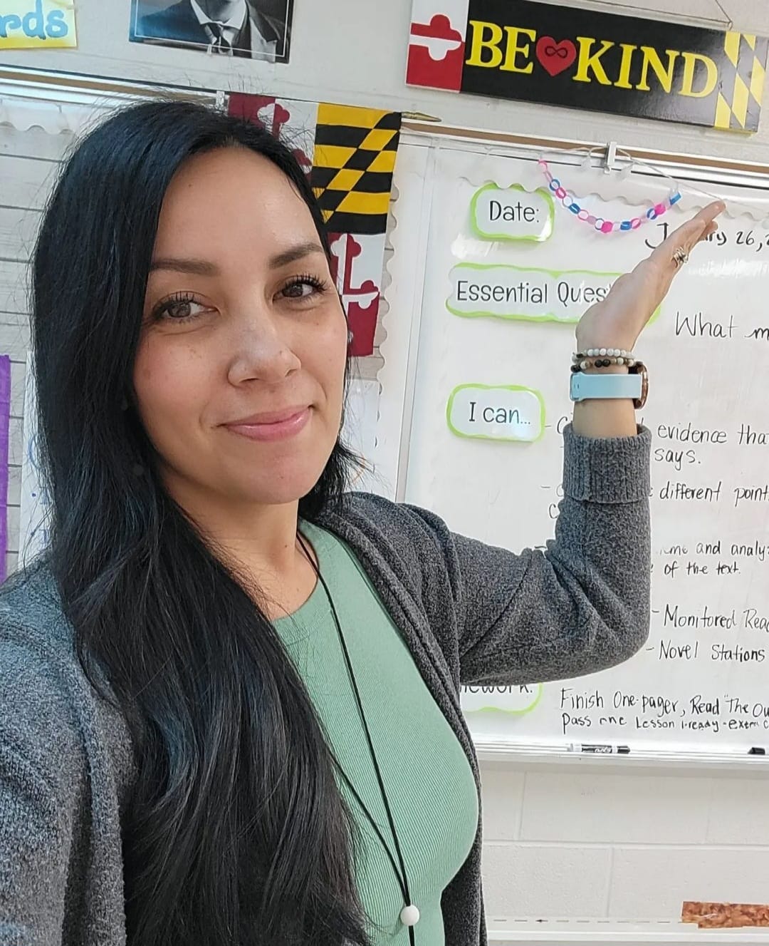 Woman with long dark hair smiling, pointing at a whiteboard with notes and decorations.