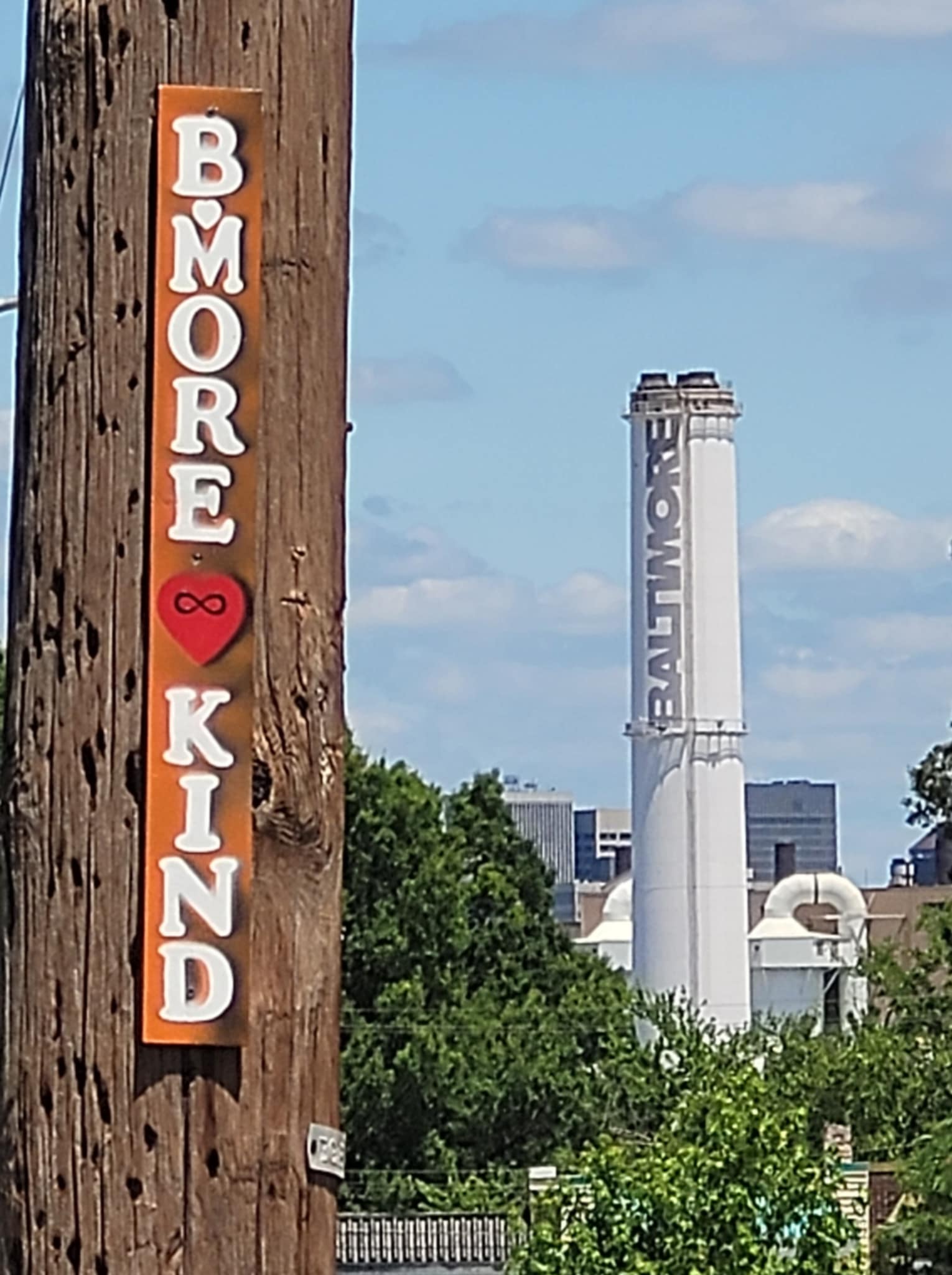 Vertical sign on a wooden post reads 'BMORE KIN' with a red heart symbol, against a cityscape background with a tall white tower.