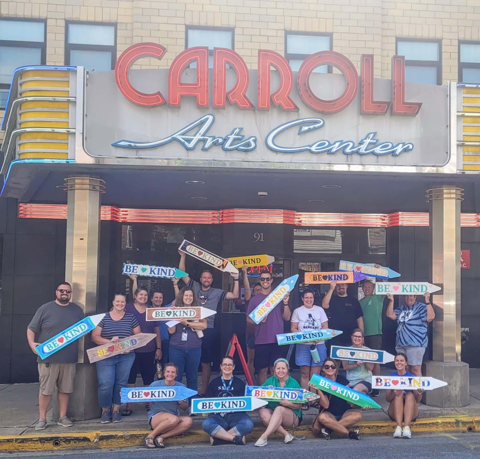 Group of people holding signs in front of Carroll Arts Center entrance, some smiling, some sitting or standing, sunny day.