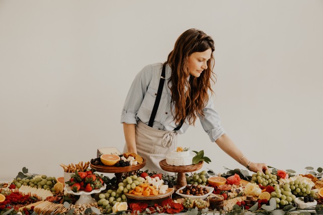Woman with long hair reaching for food on a table filled with various fruits and desserts, plain background.
