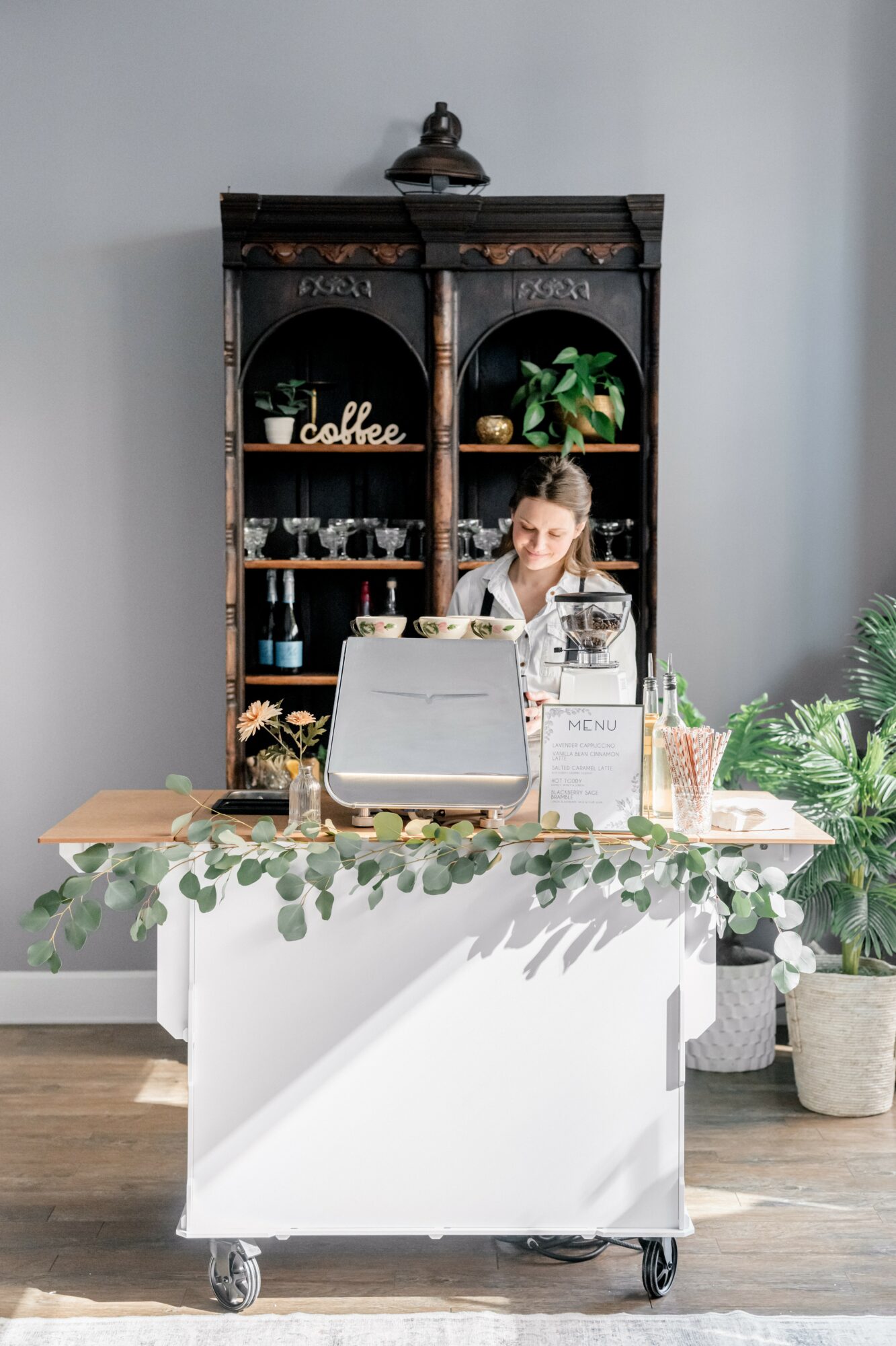 Coffee stand with a person behind the counter, shelves with coffee supplies, plants, and a white cart in front.
