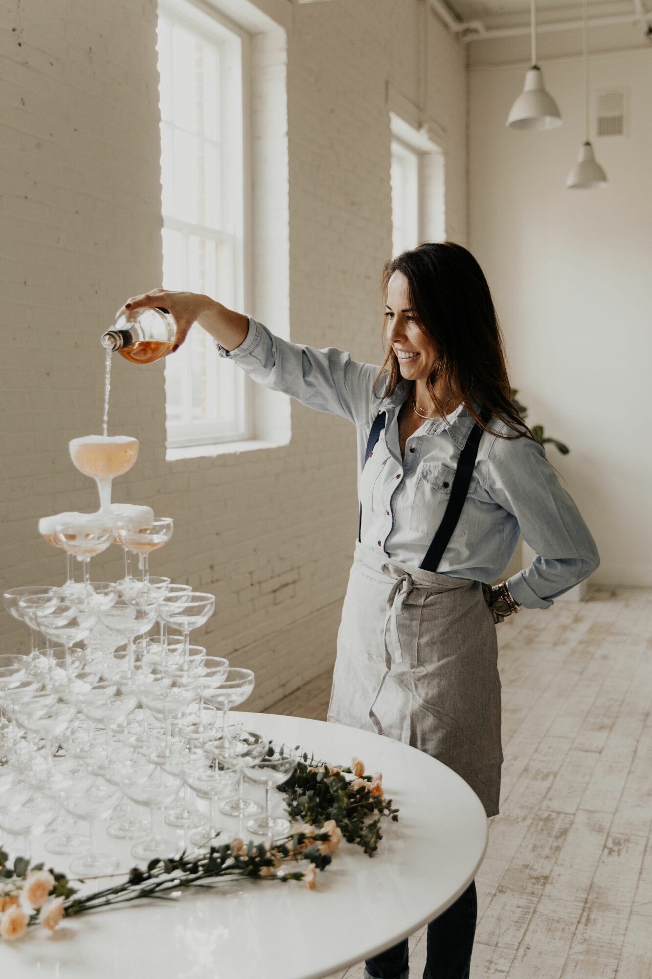 Woman pouring champagne into a tower of glasses, smiling, in a bright room with large windows and white walls.