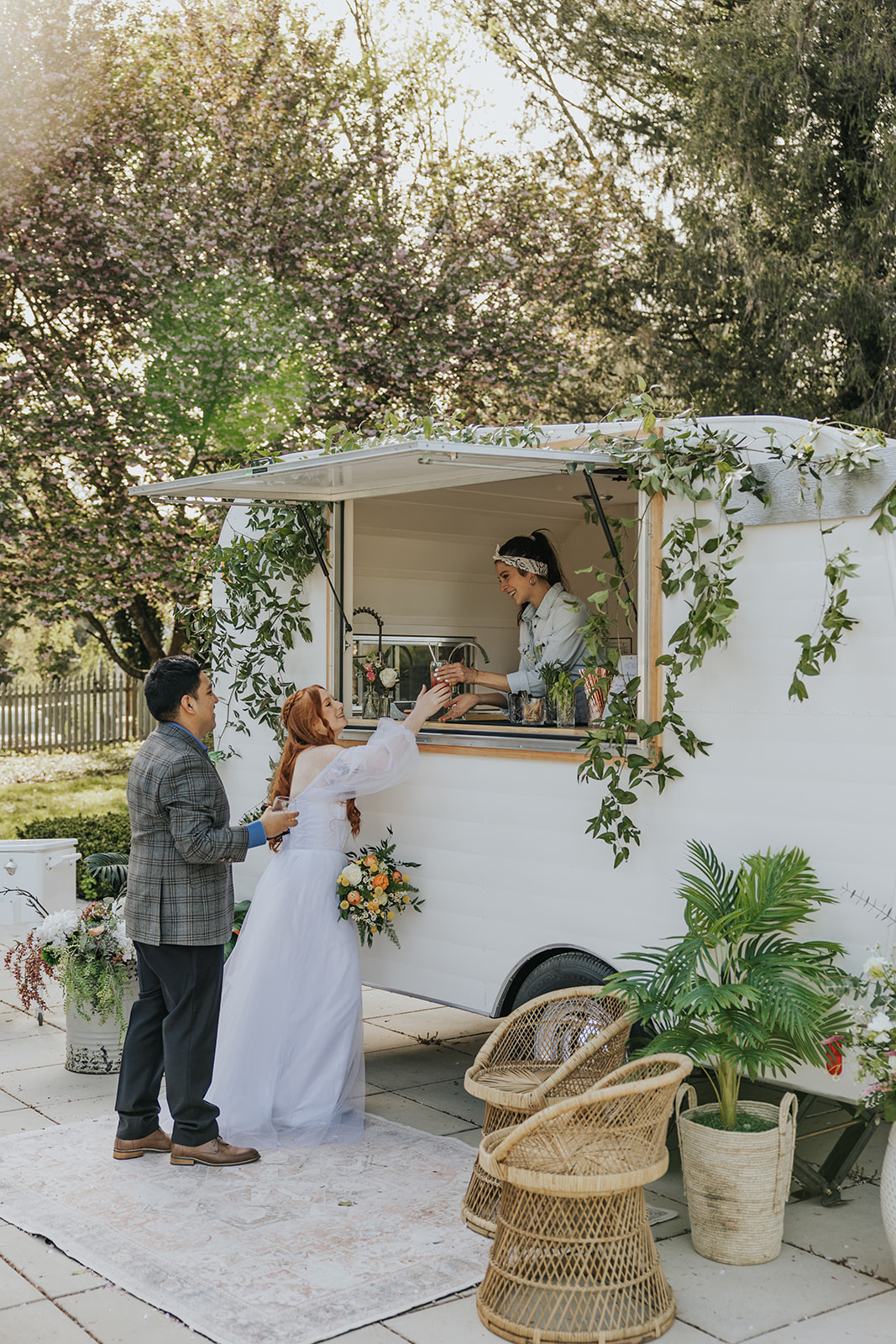 A couple at a mobile food stand, with a woman serving and a man holding a bouquet, outdoors with trees and plants.