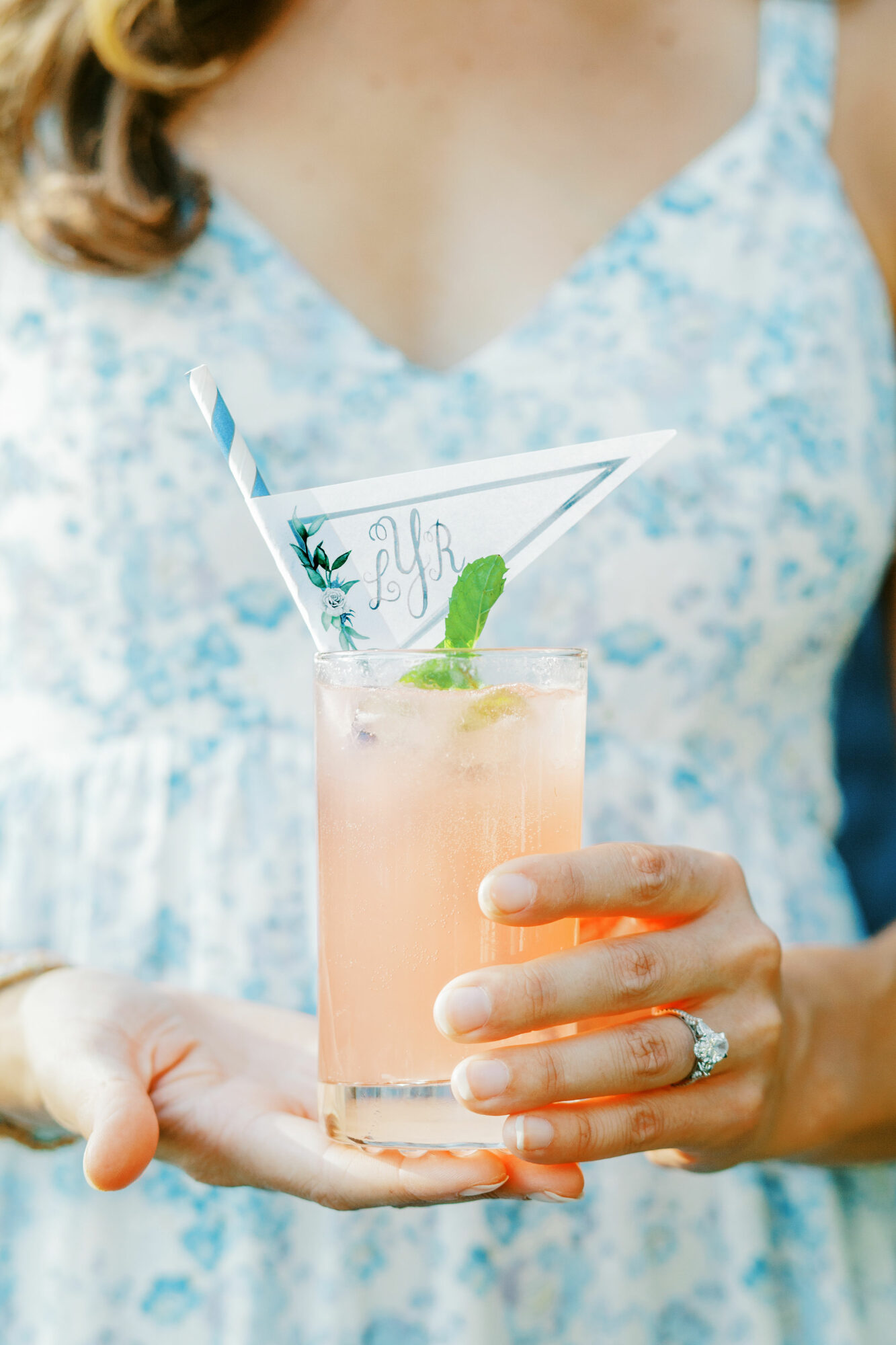 Person holding a pink cocktail with a lime wedge and decorative paper umbrella, wearing a light blue floral dress.