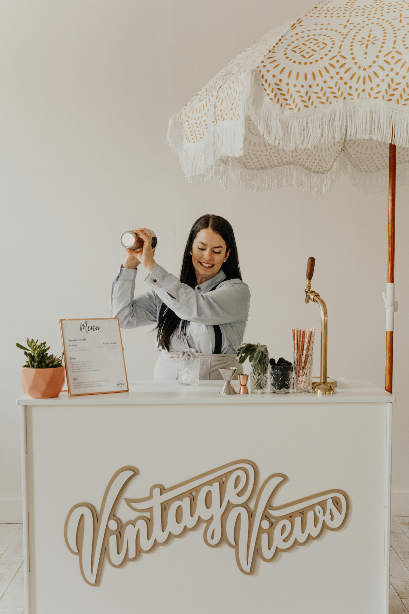 Woman smiling and shaking a cocktail shaker behind a bar with vintage decor and umbrella, in a bright setting.