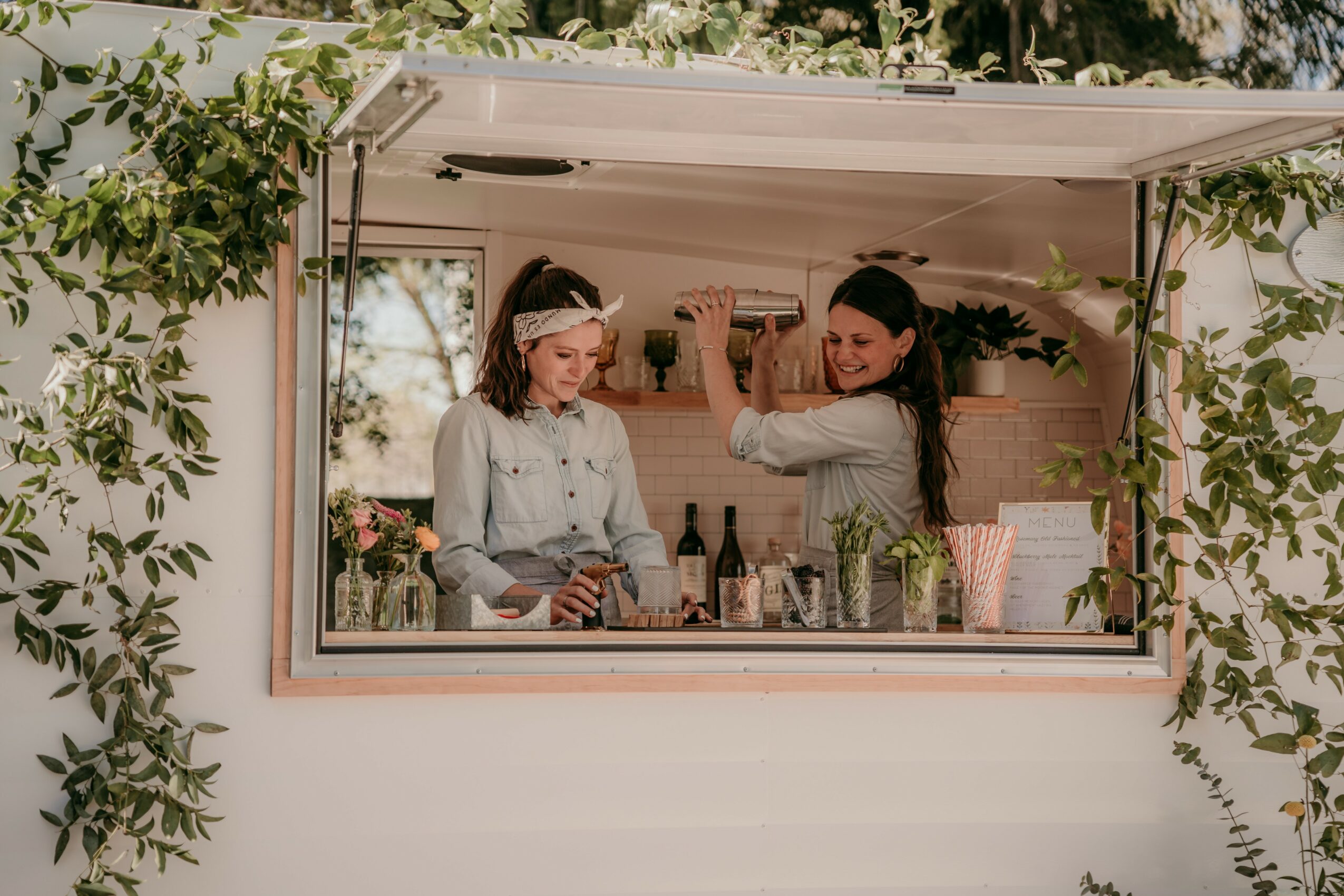 Two women preparing drinks inside a mobile bar with greenery outside, one pouring a drink, the other looking down.