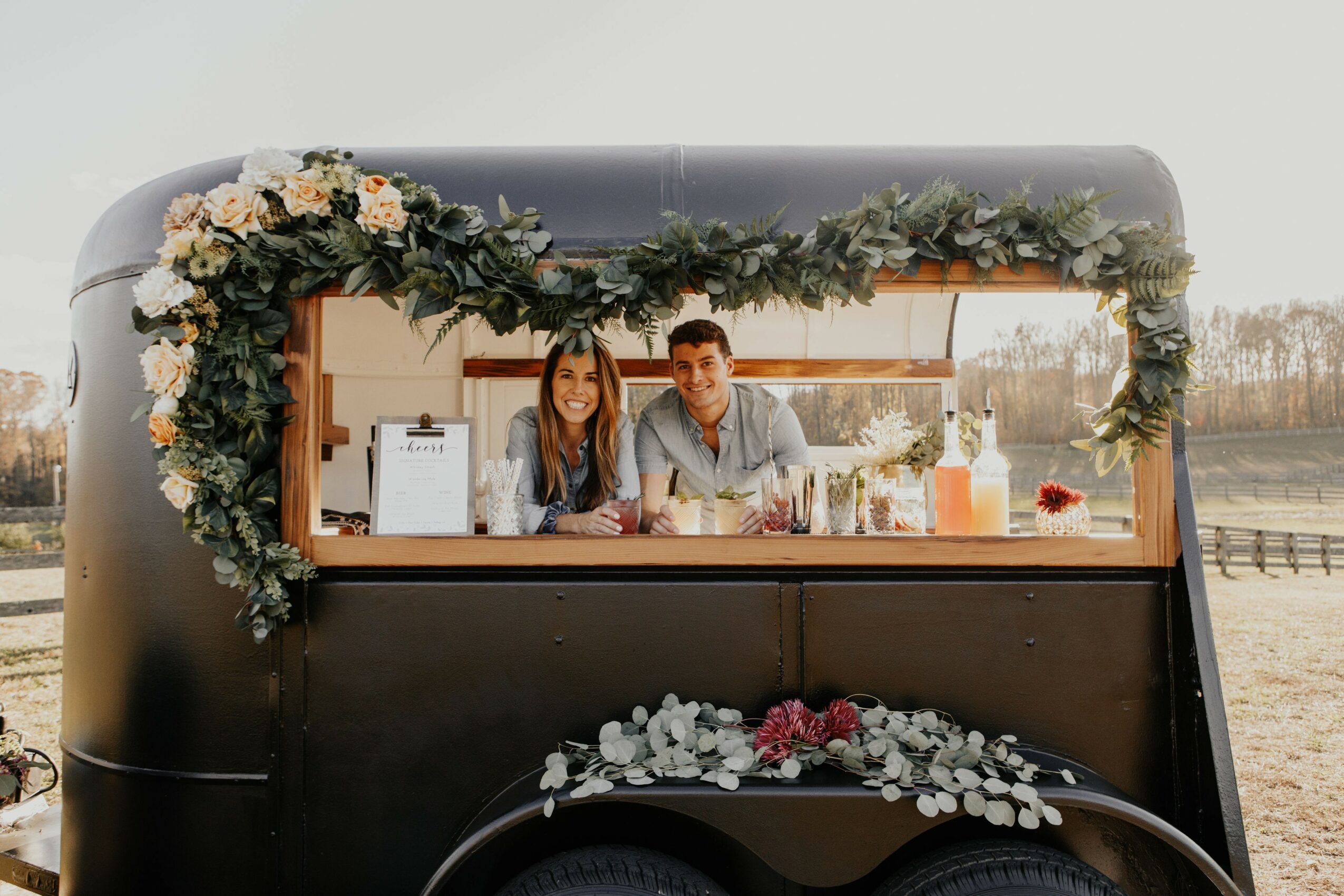 Two people sit inside a decorated food truck with flowers and greenery, looking out through the window.