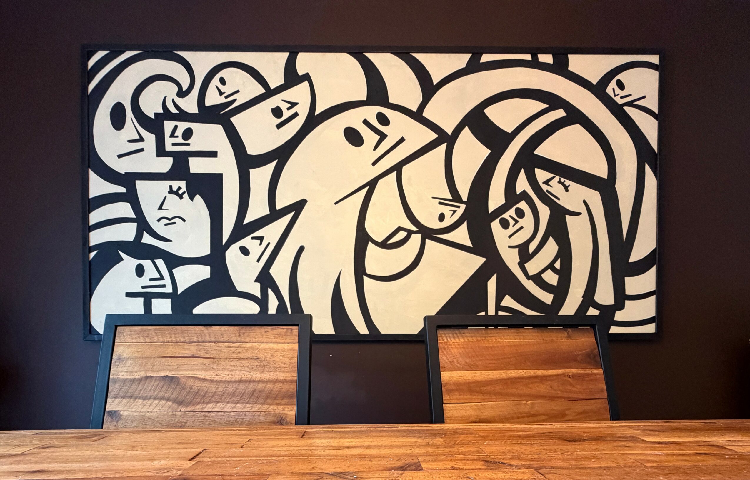 Abstract black and white artwork with various stylized faces and shapes, hanging on a dark wall above a wooden table and chairs.