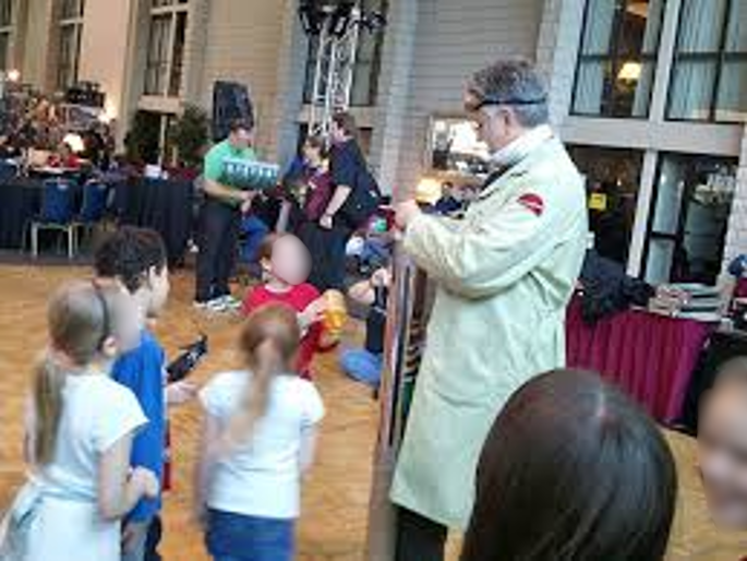 Group of children and adults in an indoor event space with tables and large windows, some children holding objects, a man in a white coat nearby.