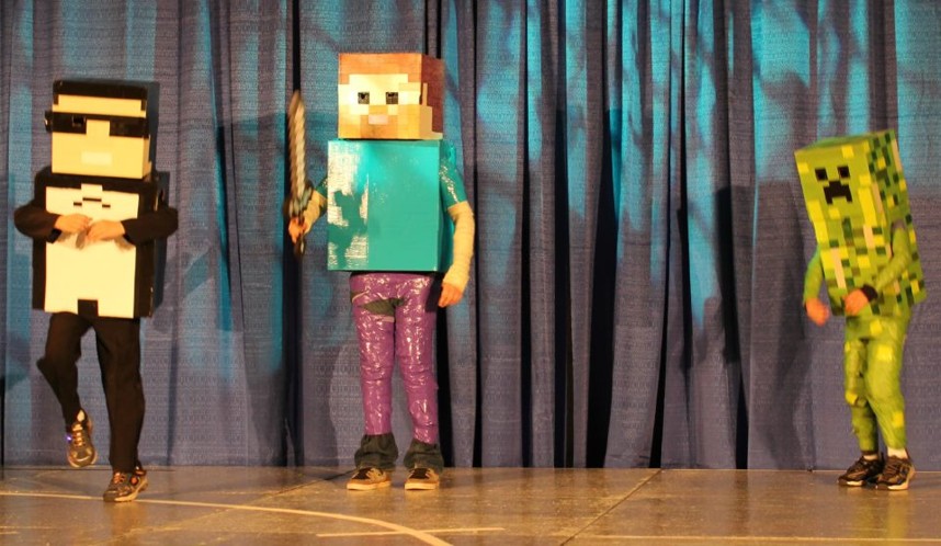 Three people in costumes on stage with blue curtain background, one with a sword, one in a pixelated outfit, and one in a Creeper costume.