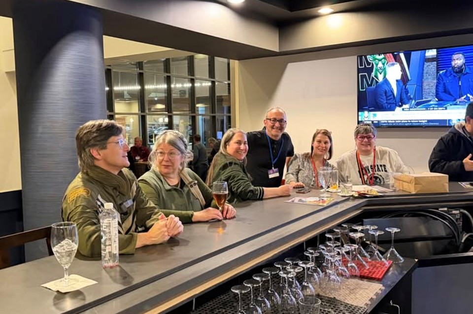 Group of seven people sitting at a bar counter with glasses and bottles, in a modern indoor setting, with a TV screen in the background.