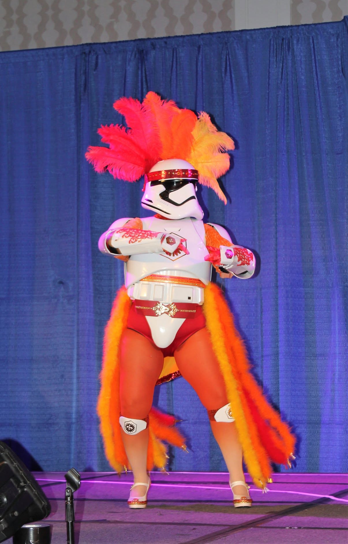 Person in a Stormtrooper costume with a colorful feathered headdress and a long orange cape, standing on stage with a blue curtain background.