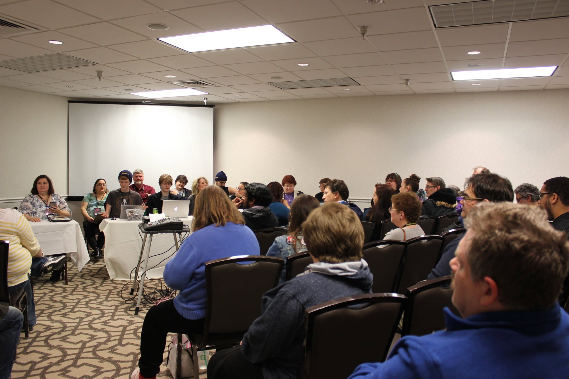 People seated in a conference room listening to a speaker at a panel, with a white wall and ceiling lights.