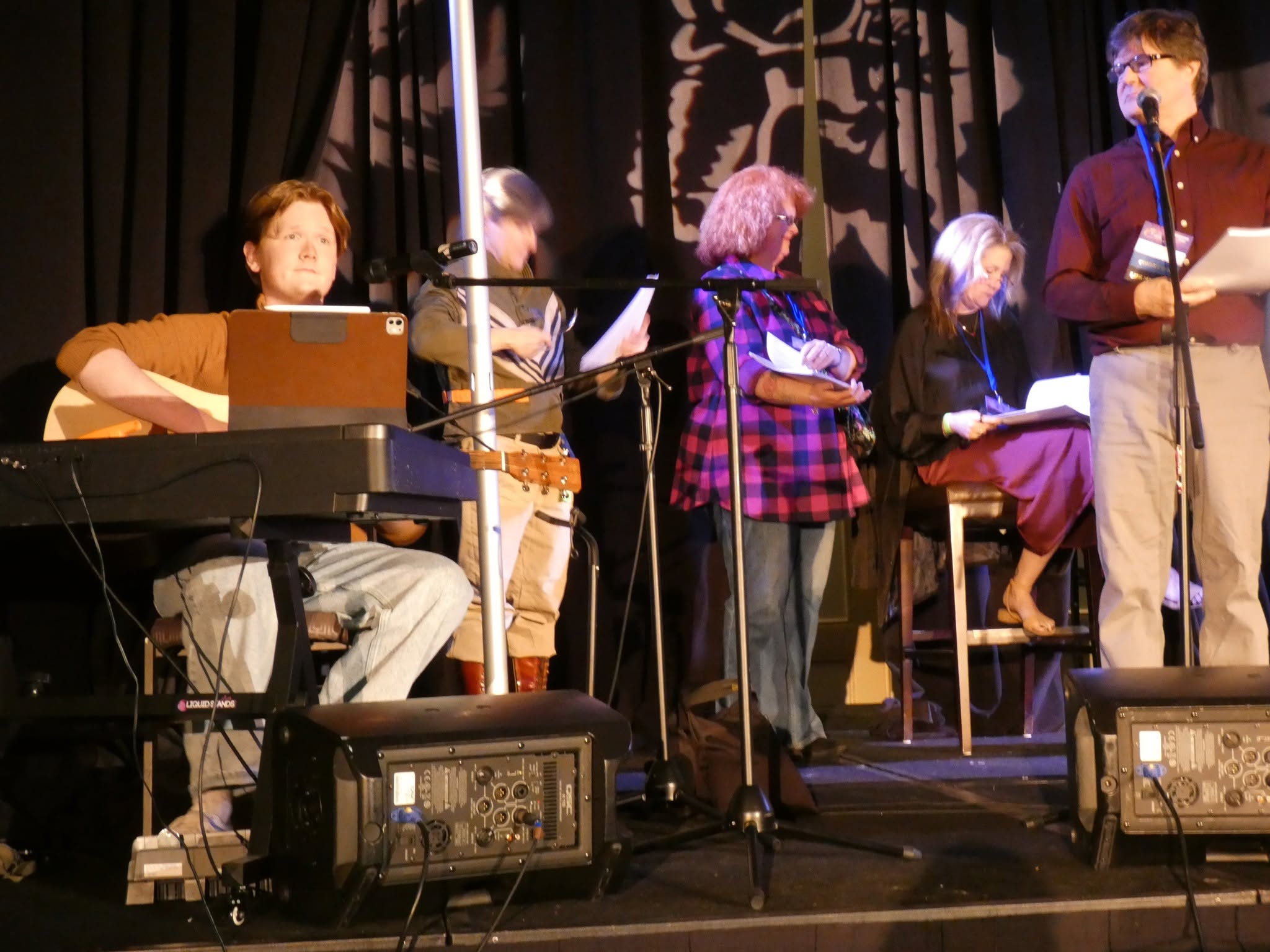 Group of people performing on stage with musical instruments and microphones, black curtain background, stage equipment visible.
