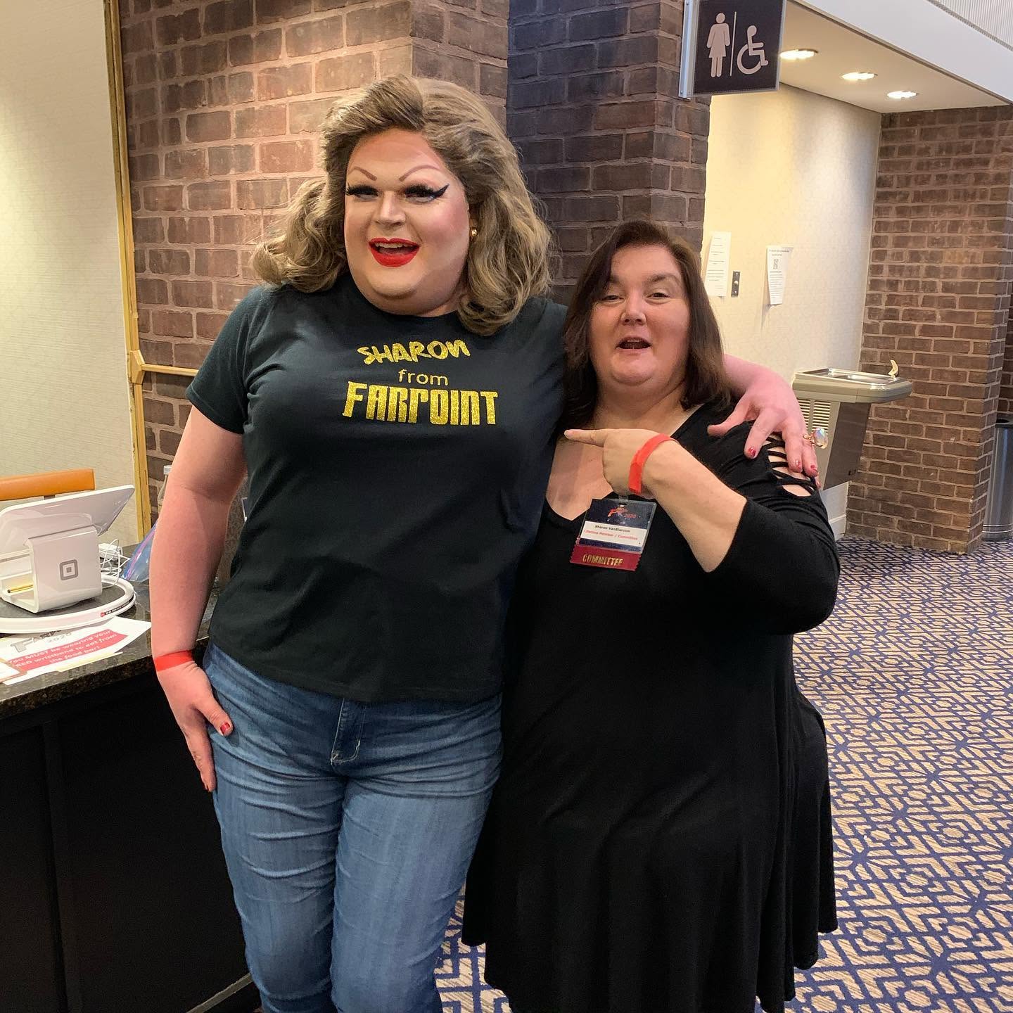 Two women standing together indoors, one wearing a black T-shirt with yellow text, the other in a black dress, smiling.