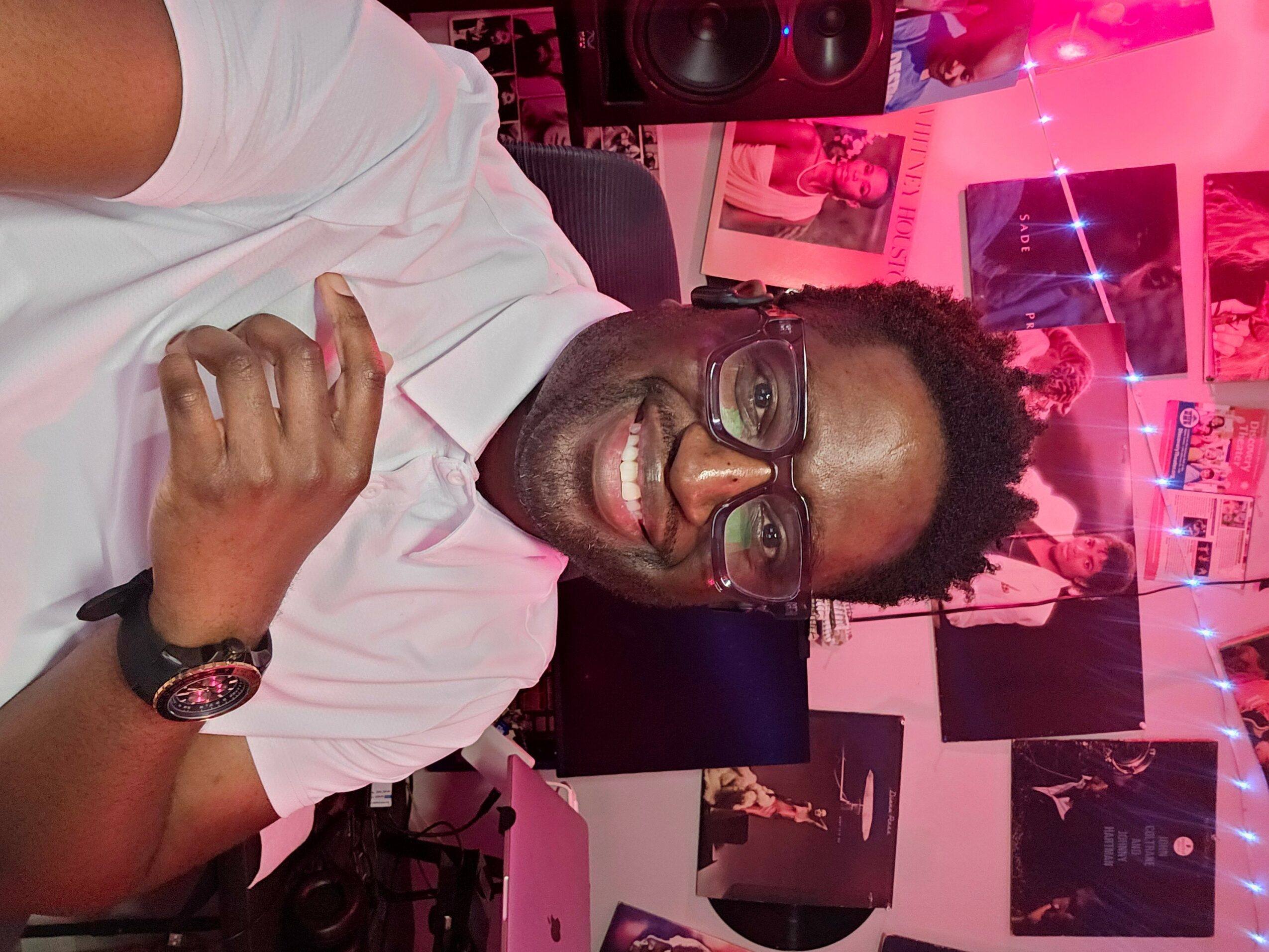 Smiling man with glasses in a room decorated with pink lighting and posters, giving a thumbs-up gesture.