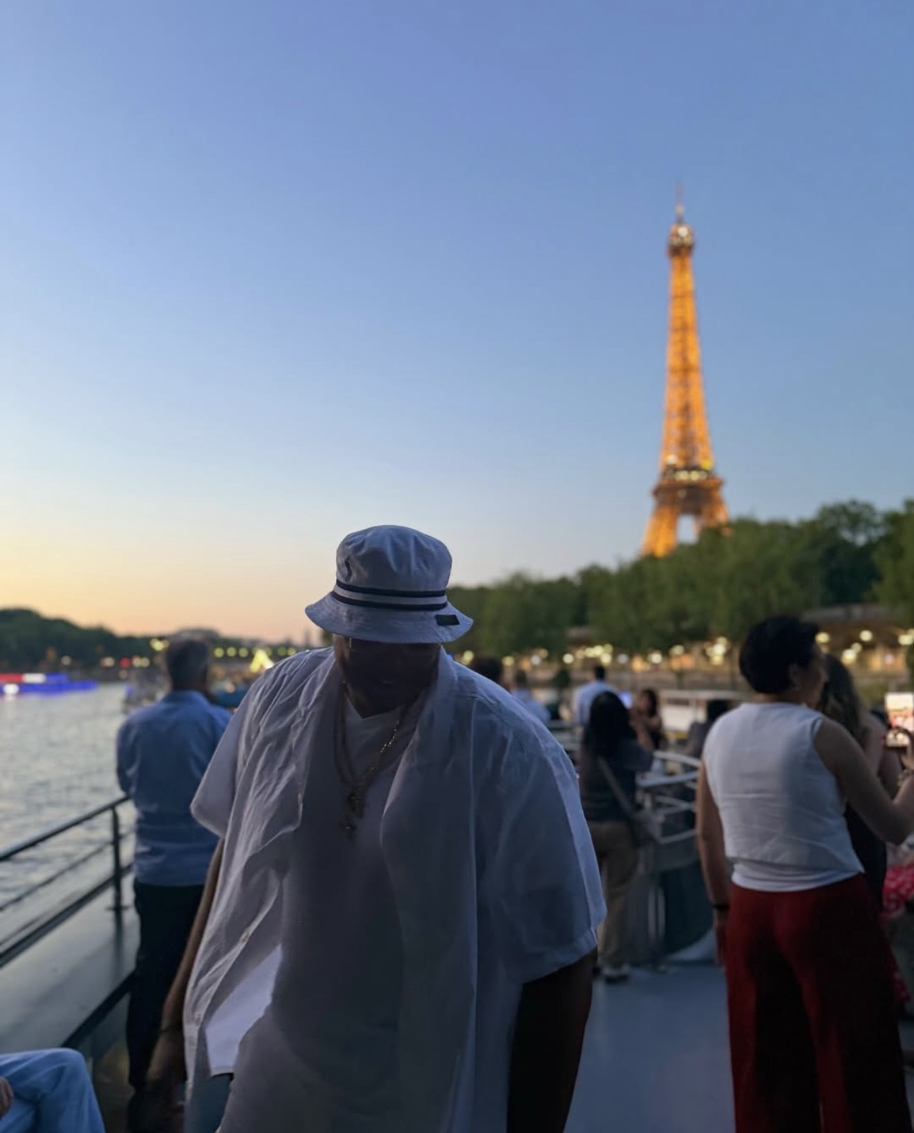 Person wearing a hat and white shirt on a boat with the Eiffel Tower in the background, during sunset.