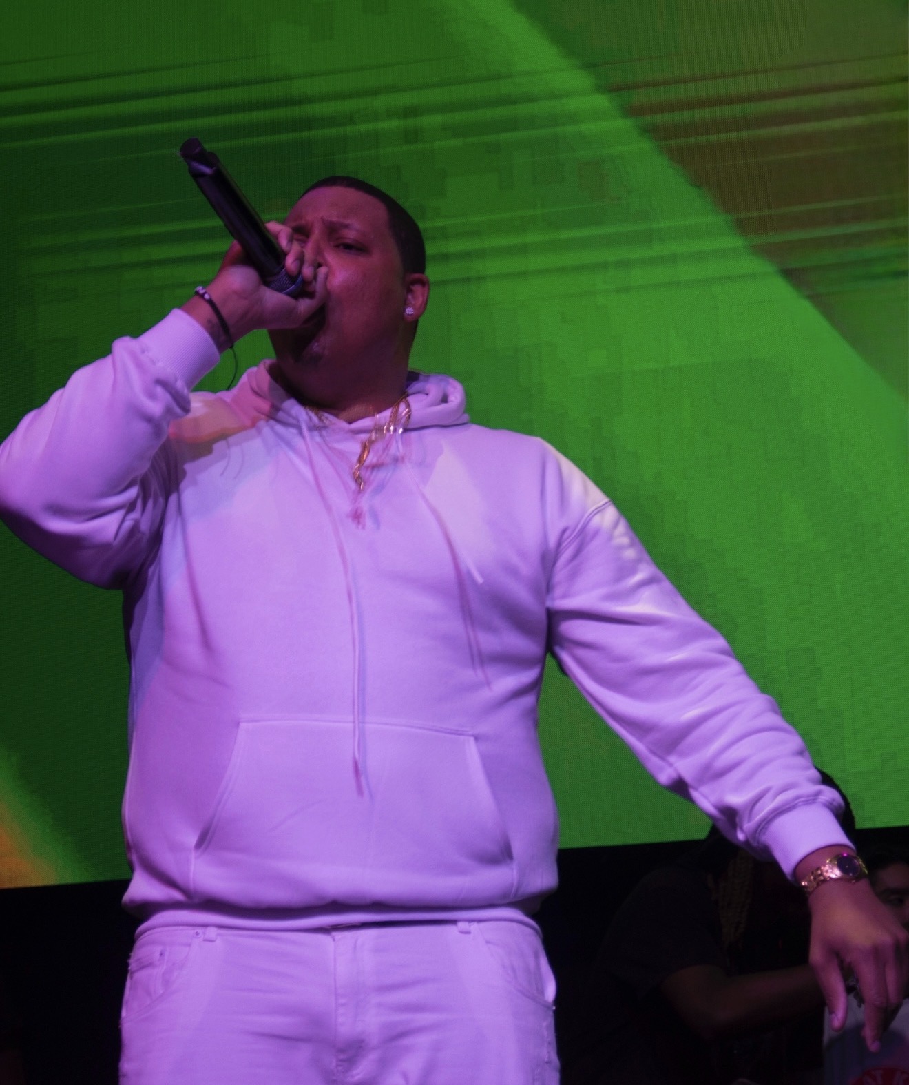 Person singing into microphone on stage, wearing a light-colored hoodie and pants, with a green background.
