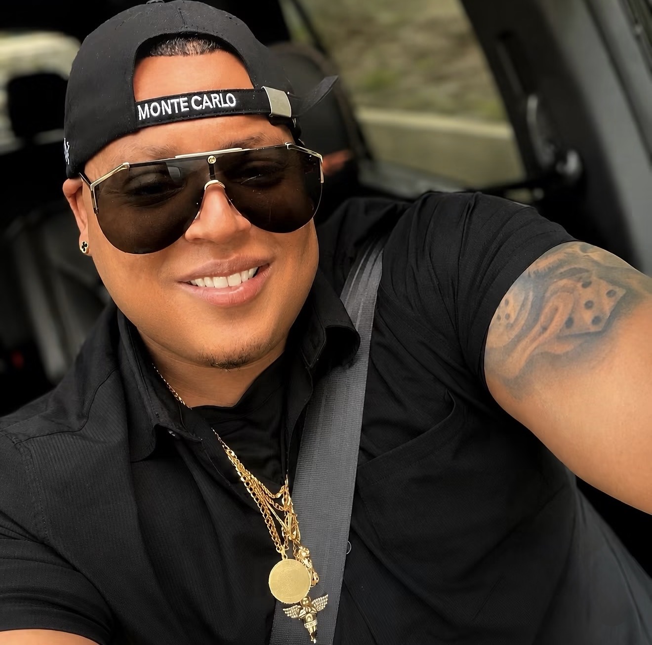 Person smiling, wearing sunglasses, a black cap, and gold jewelry, sitting in a vehicle.