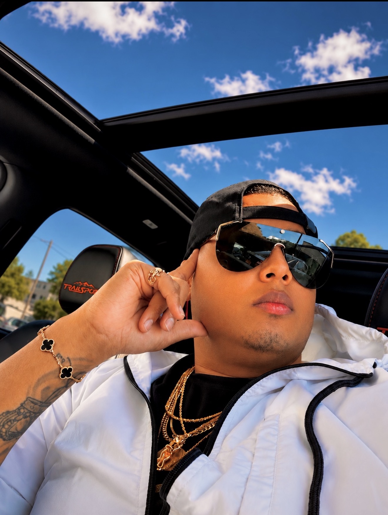 Person wearing sunglasses and a cap, sitting in a car with an open roof, against a blue sky with clouds.