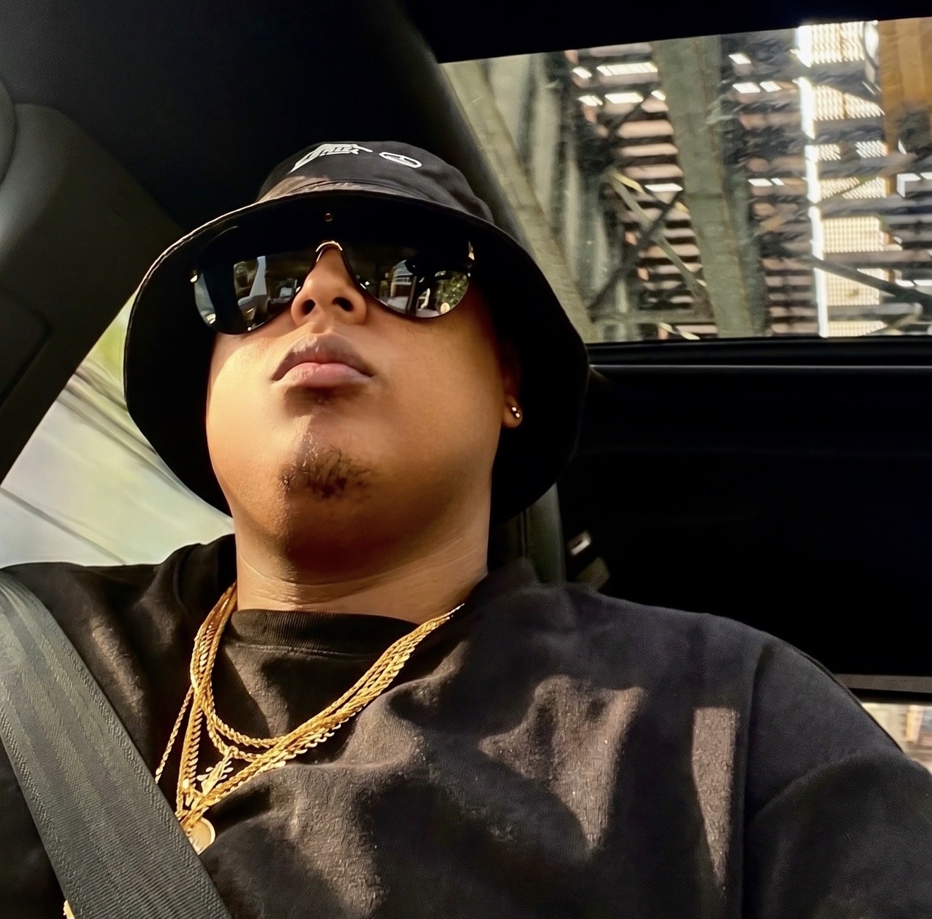 Person wearing sunglasses and a bucket hat, sitting in a vehicle with a cityscape visible through the window.