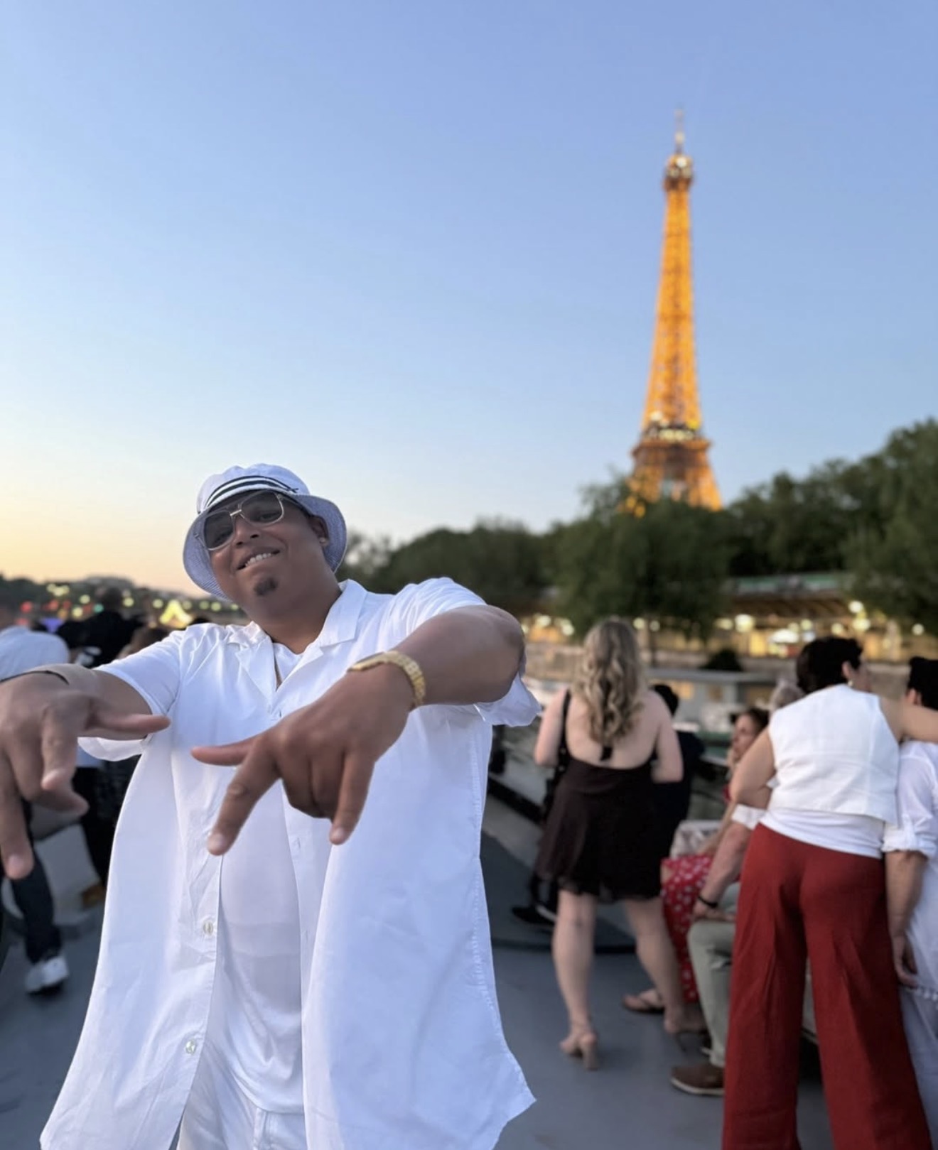 Person wearing sunglasses and a white shirt making a peace sign with hands, Eiffel Tower in background, crowd around.