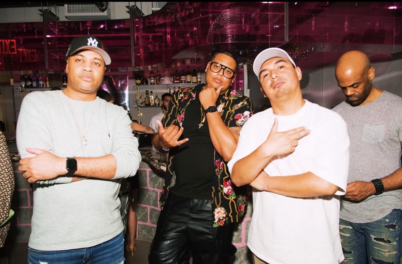 Four people standing together in a bar or club, with pink lighting and shelves of bottles in the background.
