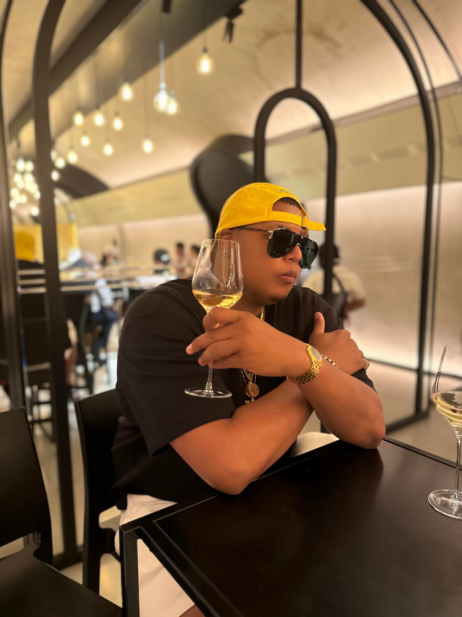 Person wearing sunglasses, yellow cap, and black shirt sitting at a table with a glass of wine in a restaurant.
