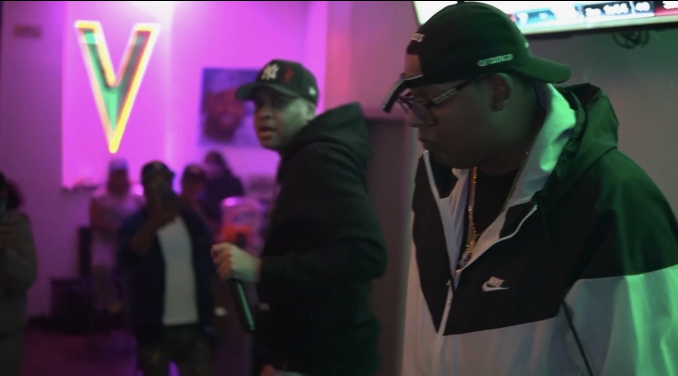 Two men in hoodies and caps in a dimly lit room with a colorful neon V sign, people in the background.