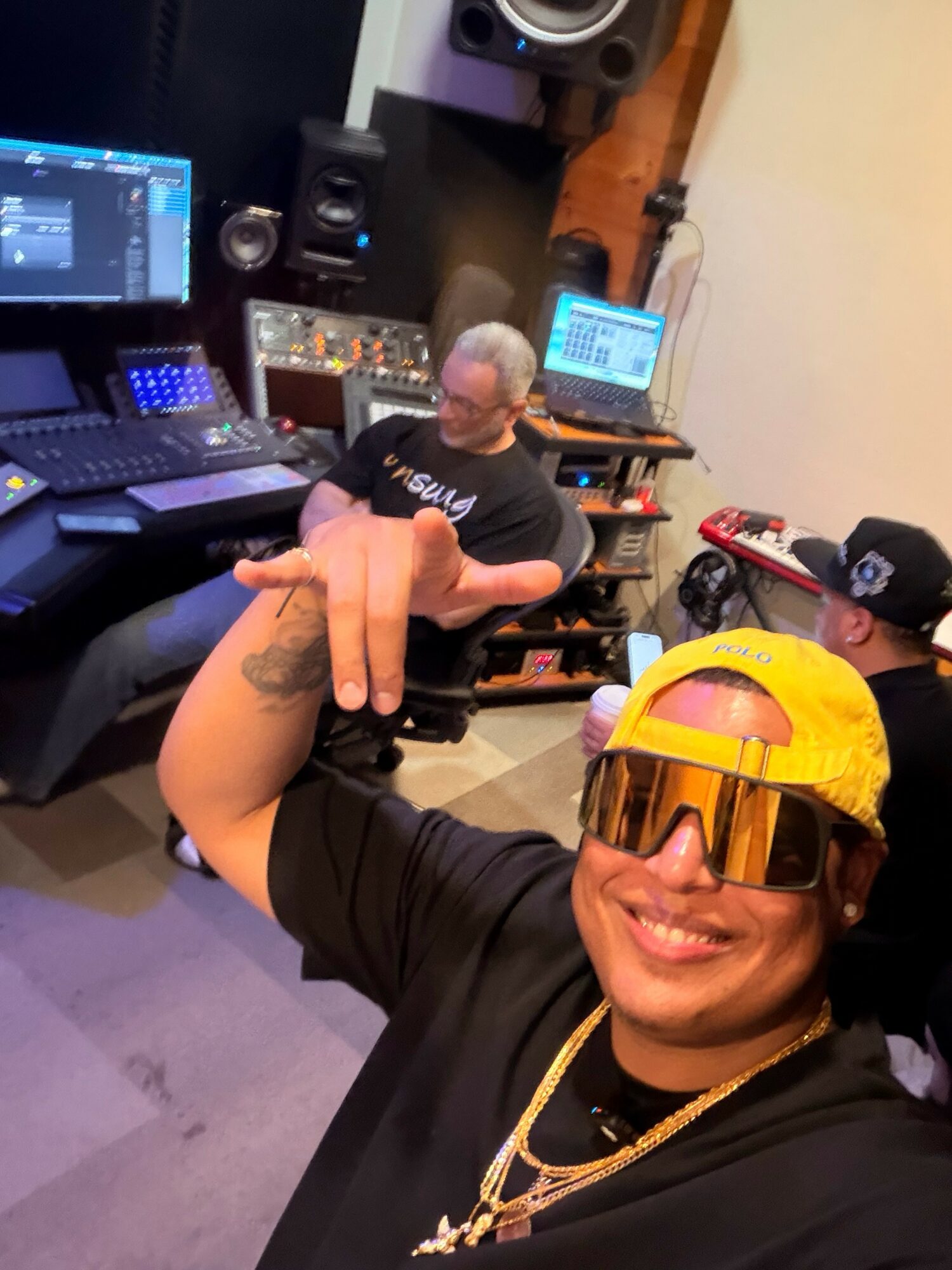 Person wearing yellow cap and sunglasses smiling, in a music studio with equipment and another person working at a computer.