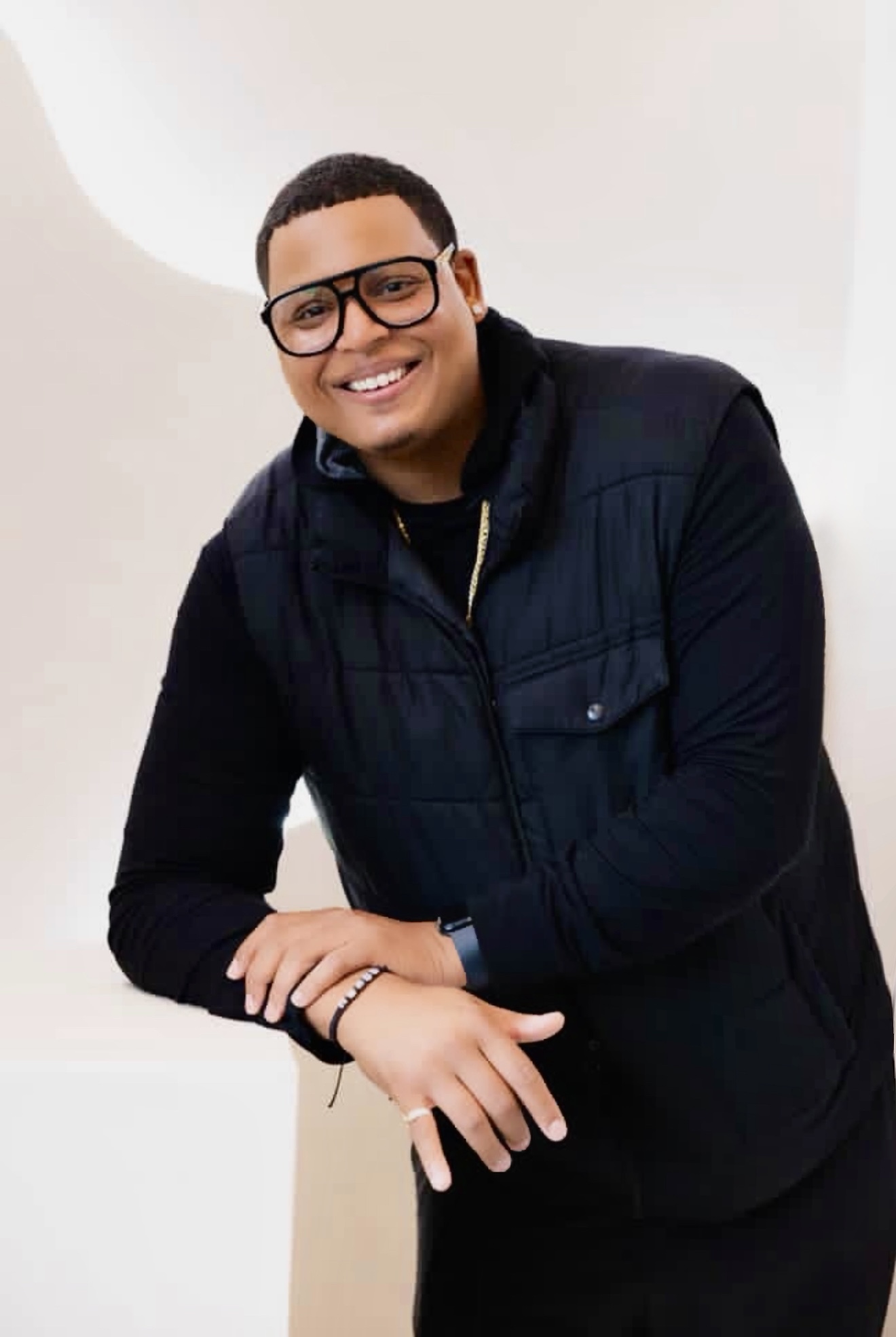 Person with glasses smiling, wearing a dark jacket, leaning on a surface, against a plain background.