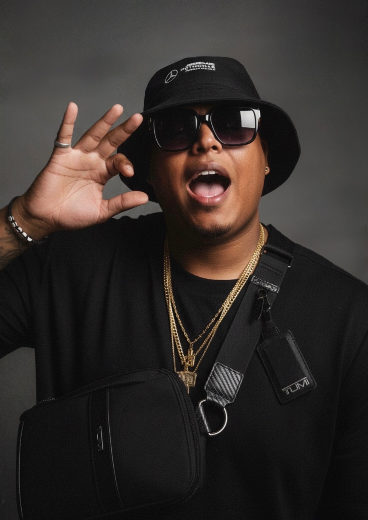 Person wearing sunglasses, bucket hat, and gold chains, holding a black bag, with mouth open and hand near ear.