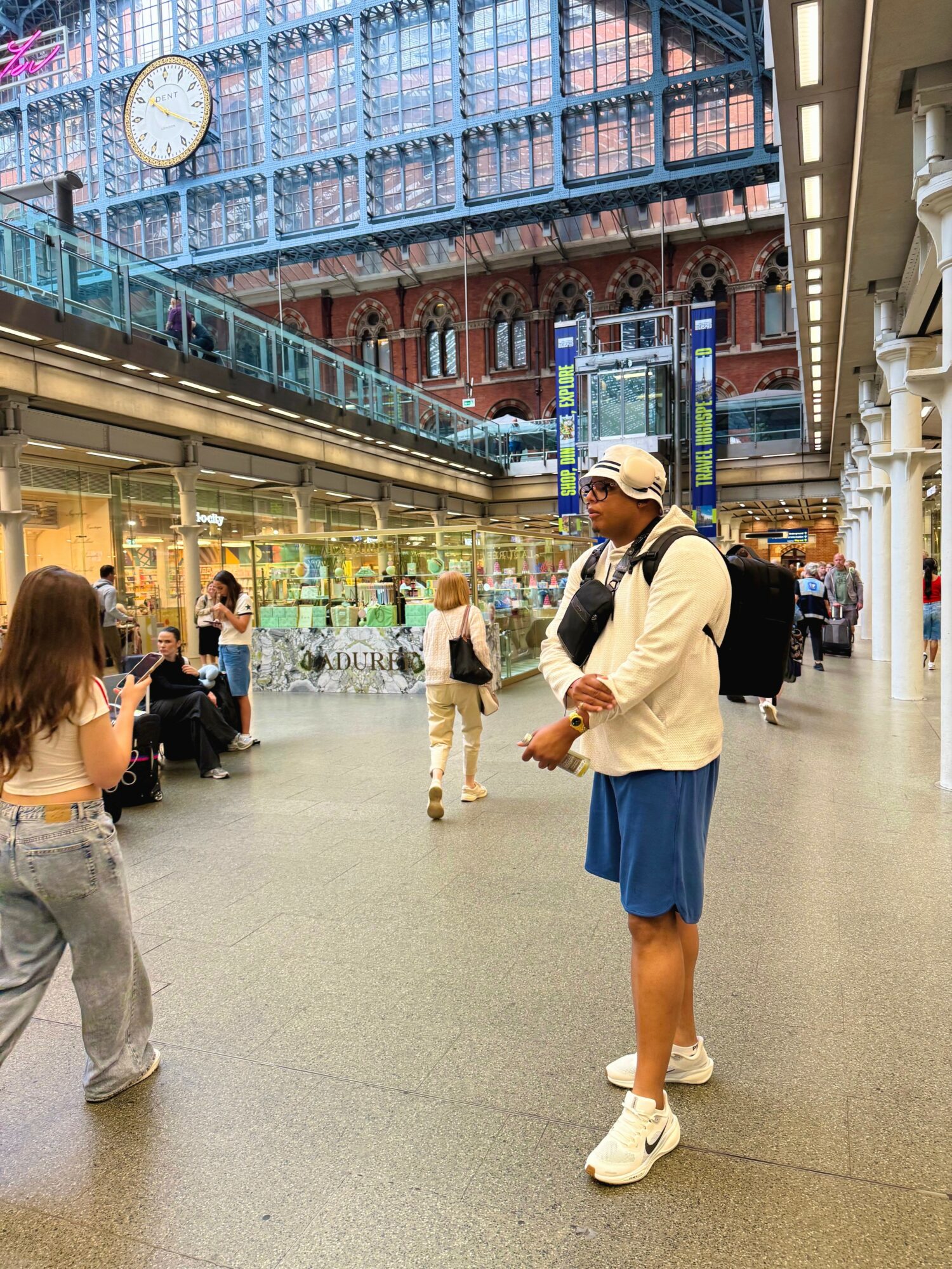 Person wearing a white jacket, blue shorts, white shoes, and a cap standing in a busy indoor shopping area with a large clock and glass ceiling.