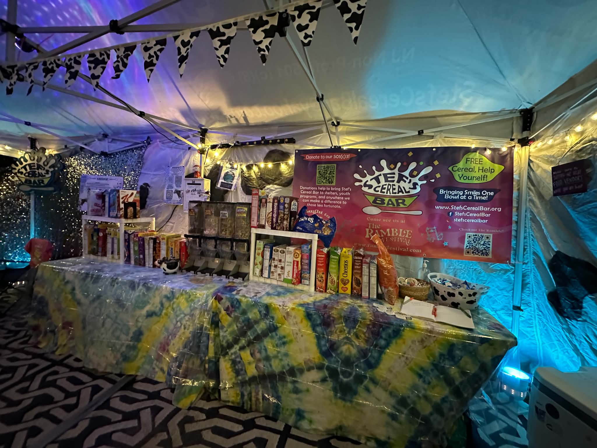 Table with books and items under a tent with colorful lighting, banners, and patterned floor, at an event or market.