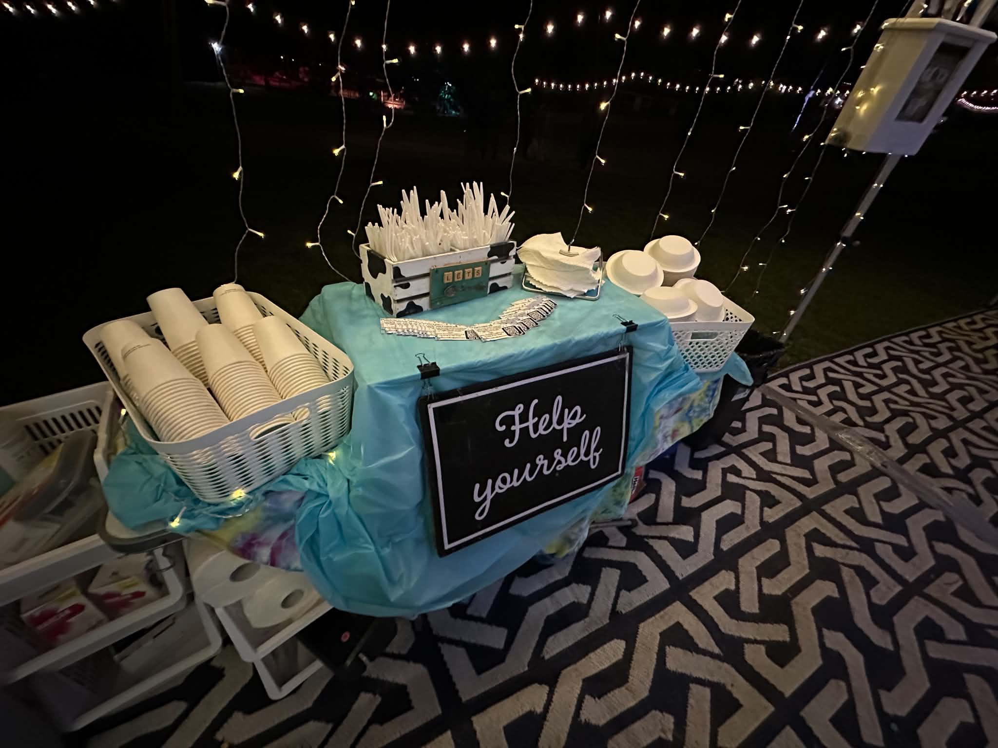 Table with napkins, plates, and decorations, with a sign saying 'Help yourself,' under string lights, on a patterned floor.