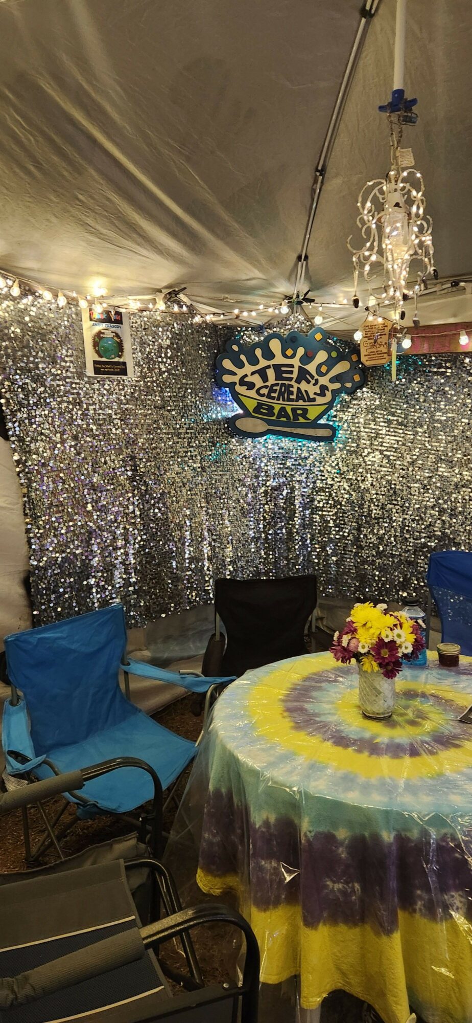 Party table with a yellow and purple cake, flowers, and a glittery backdrop, with chairs and a person in blue nearby.