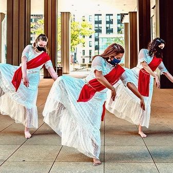 Three women in white dresses with red sashes perform a dance indoors, wearing face masks, with large windows and city buildings in the background.