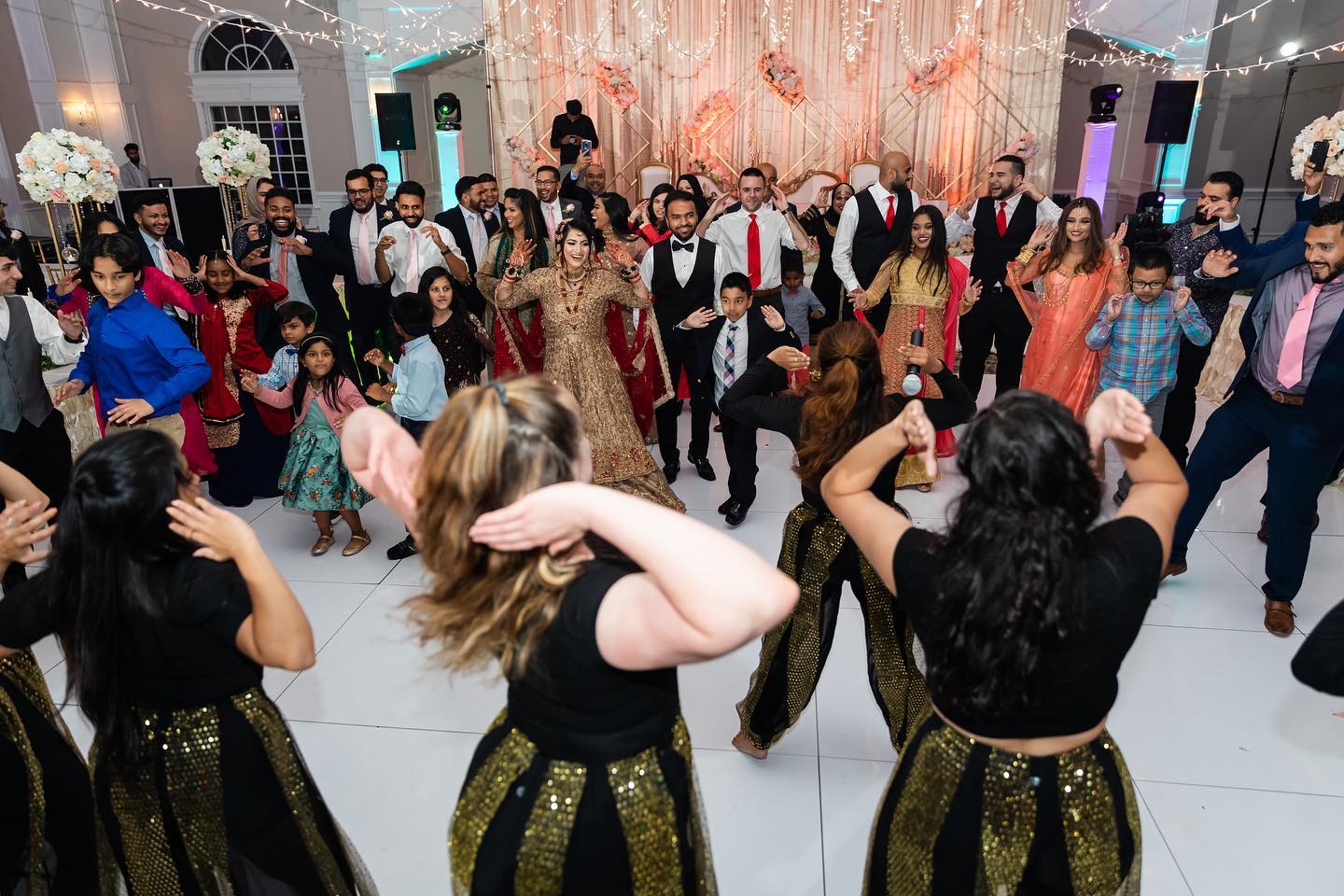 People dancing at a celebration in a decorated hall with a pink backdrop, some wearing traditional attire, others in formal wear.