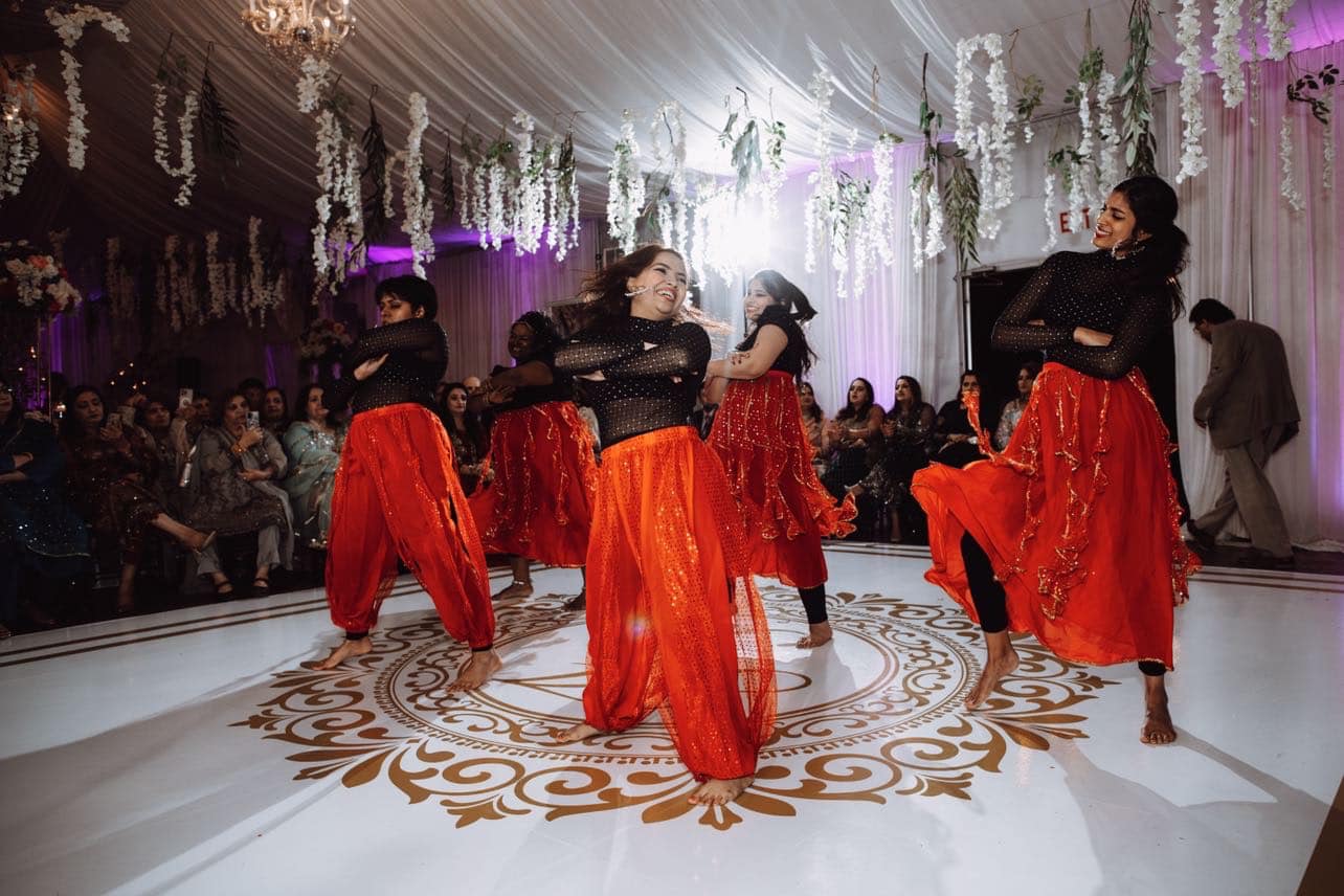 Four dancers in black tops and red skirts perform on a decorated stage with an audience watching.