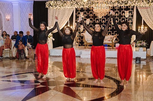 Four women dance in black tops and red pants on a decorated stage, with an audience in the background.