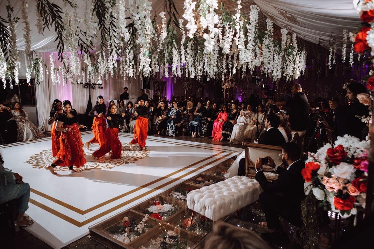 Group of dancers performing on a decorated stage with hanging flowers and an audience watching, in an indoor event space.