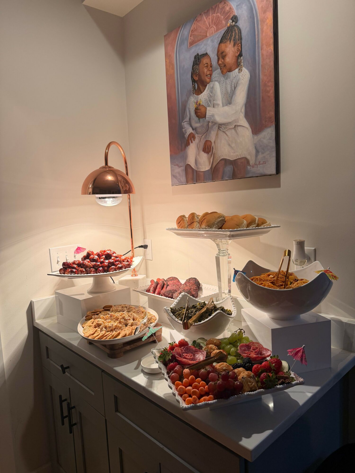 Table with desserts, fruit, and a cake, with a family portrait on the wall behind. Bright lighting and a white table.