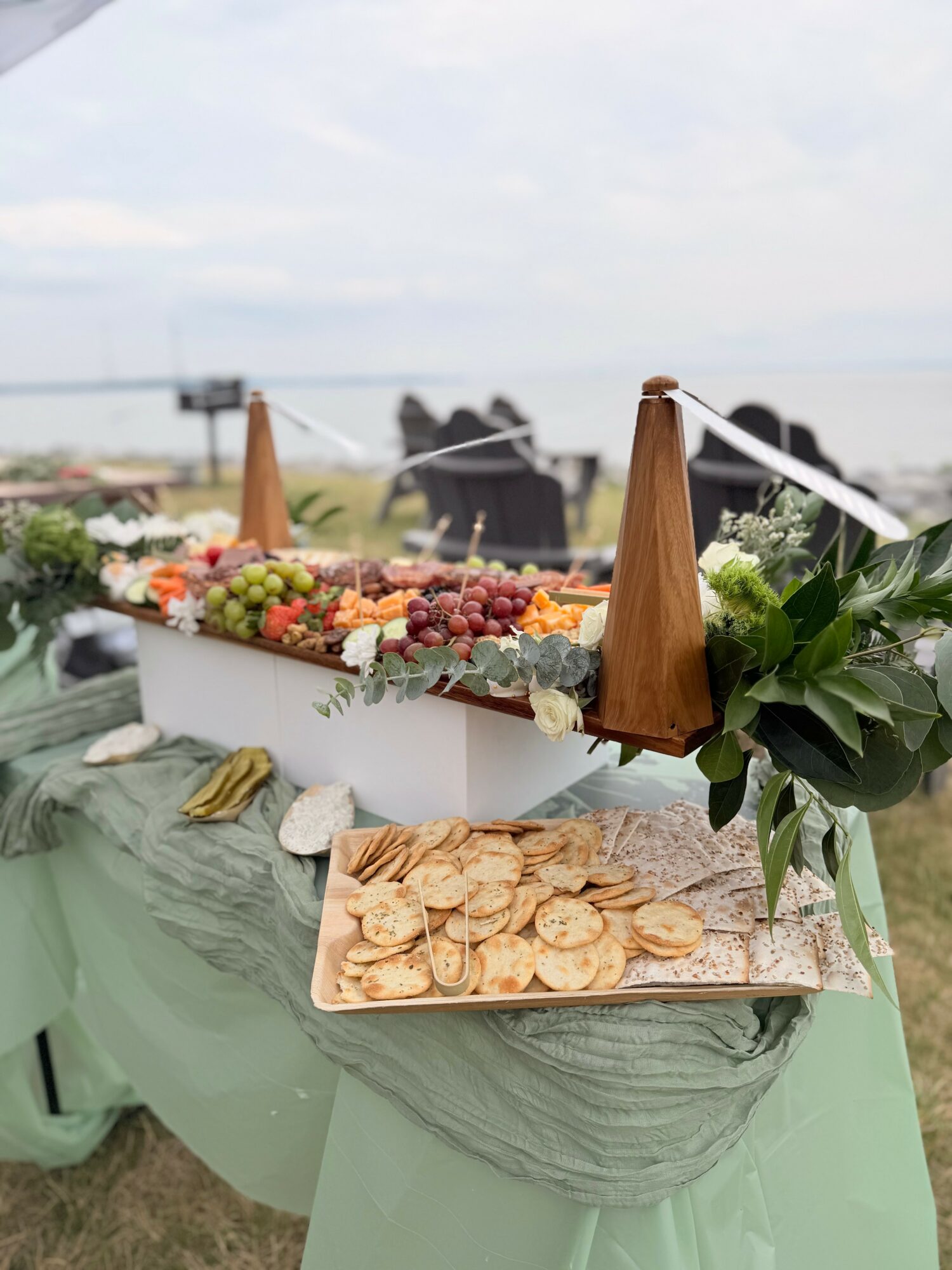 Table with fruit and snack platter outdoors, with a lake and horses in the background, under a cloudy sky.